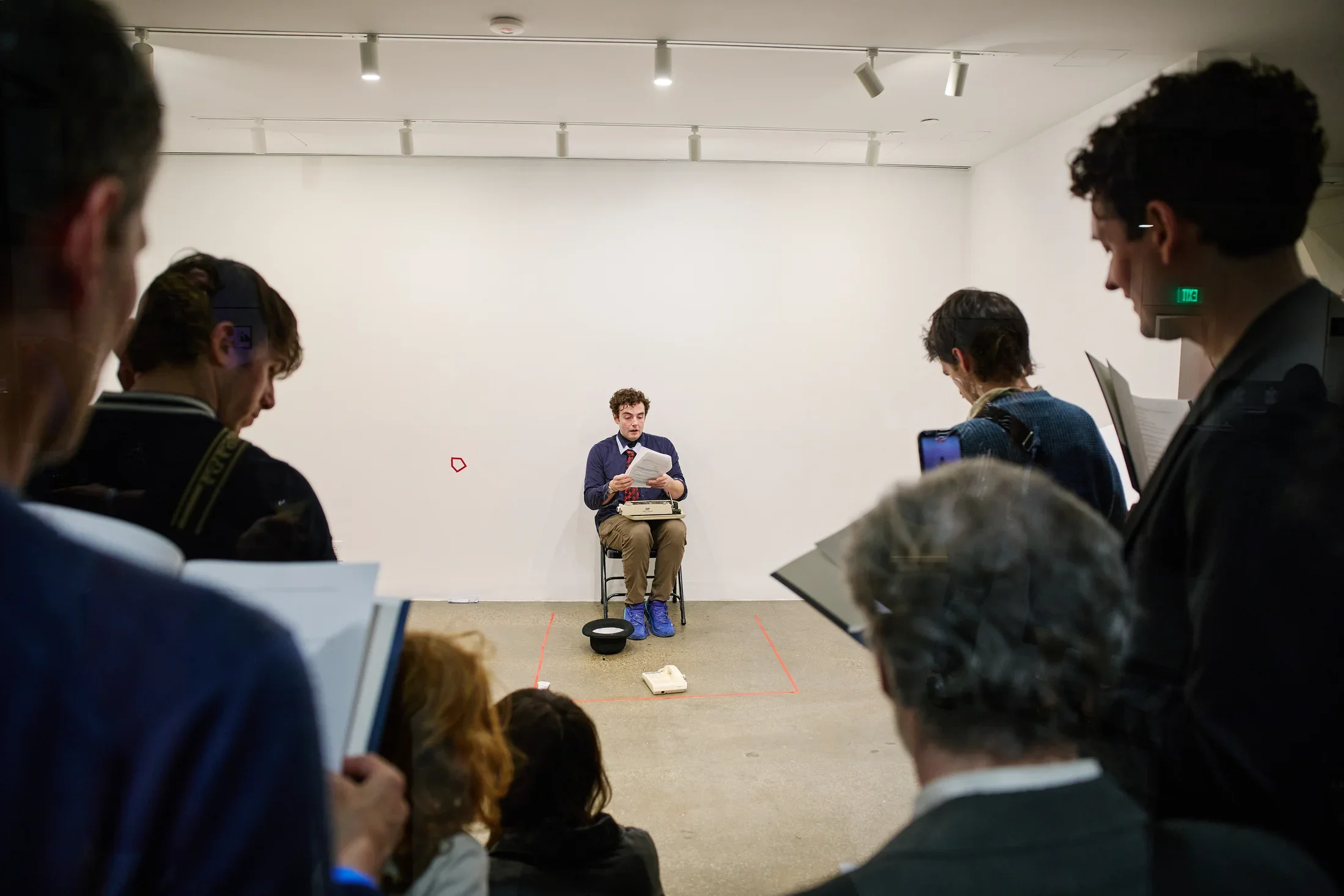 An actor sits on a metal folding chair and reads from a script. A typewriter sits in his lap. We glimpse him over the shoulder of several onlookers, who refer to copies of the script in folders.