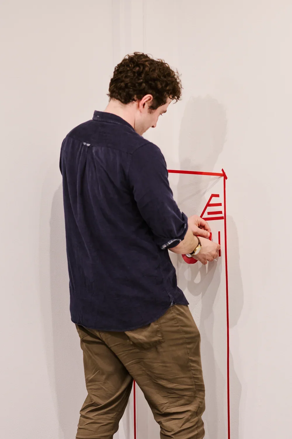 A man stands in front of a corner with white walls, adding thin lengths of red tape to the walls. They connect to form an abstract geometric design.