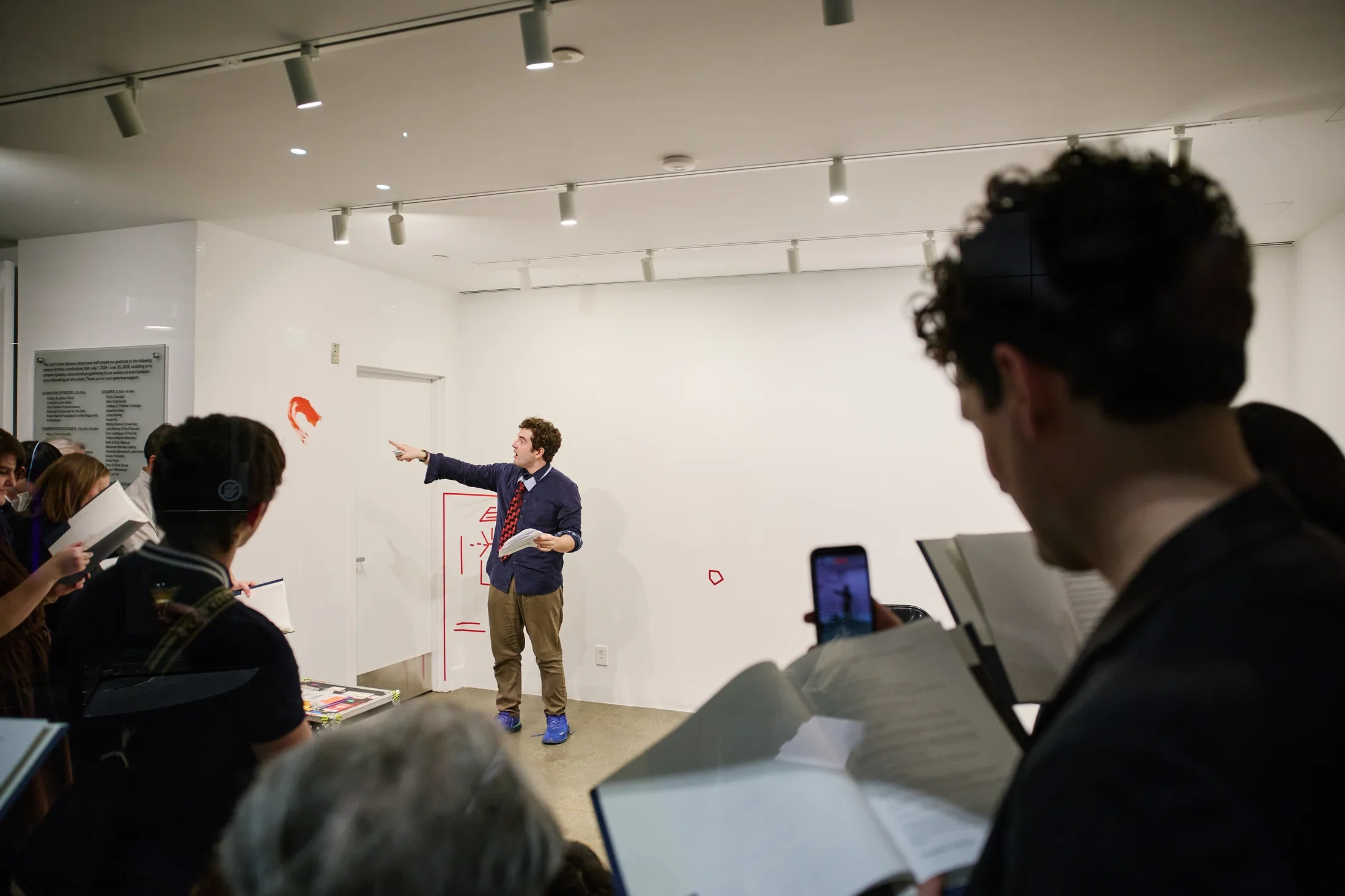 An actor holds a printed script and points to a smear of red paint on the wall of a gallery. Behind him, the wall is marked by some abstract shapes created by colored tape. We glimpse him over the shoulder of several onlookers, who refer to copies of a script in folders.