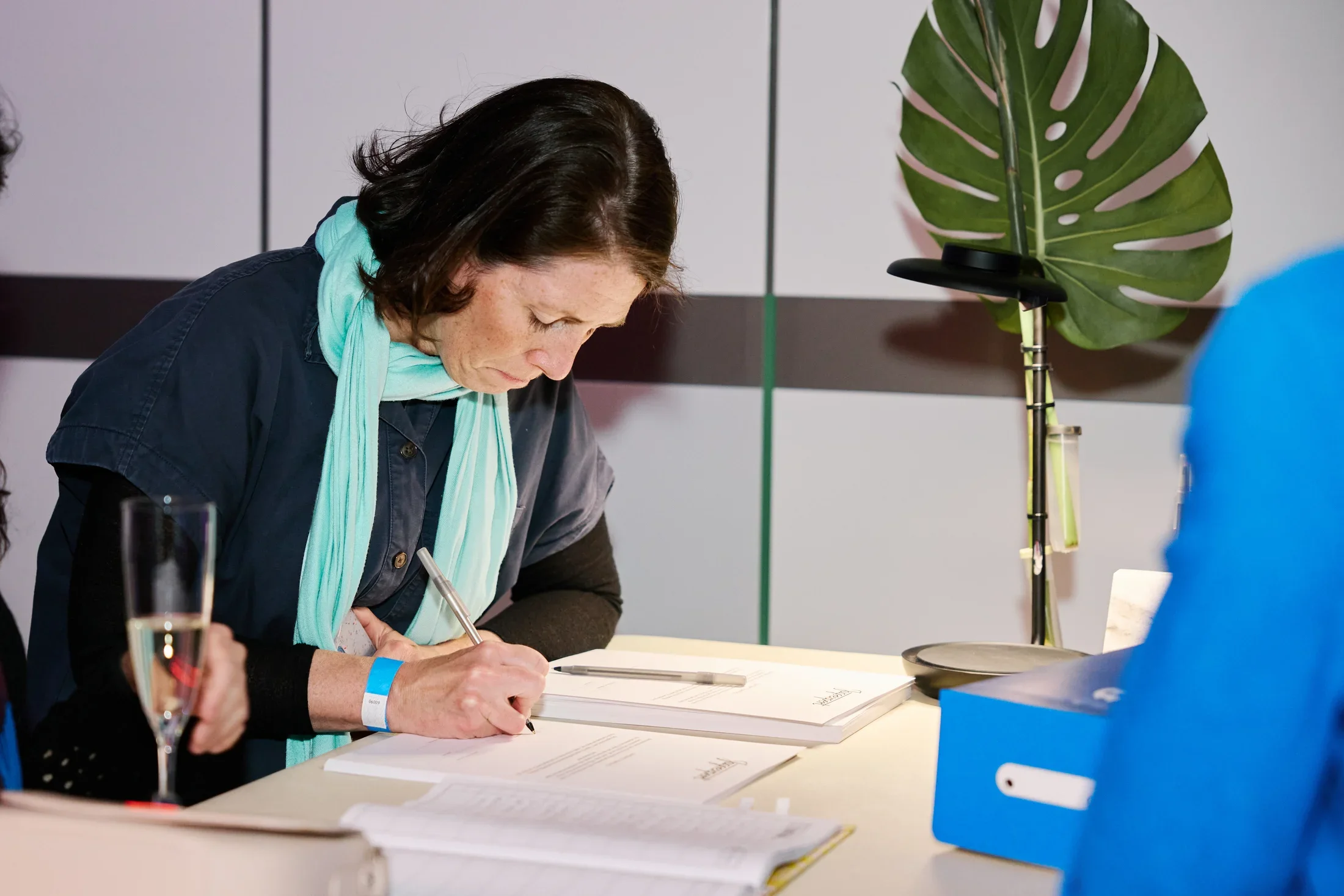 A woman wearing a pale aqua scarf leans over a table and uses a pen to sign a contract. Other contracts and papers are visible on the table, along with a flute of wine, a blue shoebox, and a lamp with a monstera leaf attached.