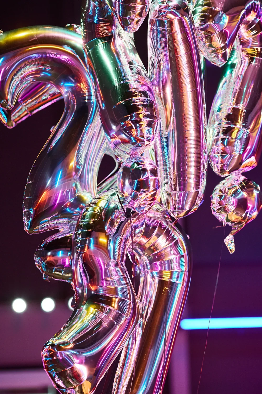 A cluster of shiny, iridescent silver balloons shaped like letters and punctuation marks, including two question marks, reflects pink, purple, and blue light against a dark, blurred background.