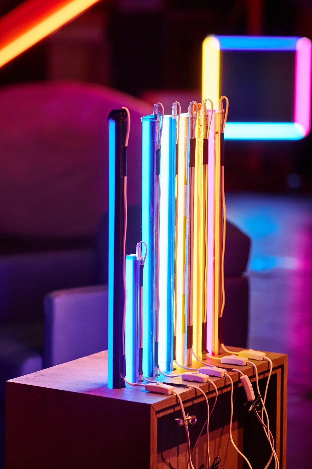 A row of vertical fluorescent light tubes, glowing blue, pink, and orange, stands on a wooden box. Wires with switches connect to the bases of the tubes, and additional colored lights are visible in the dark background.