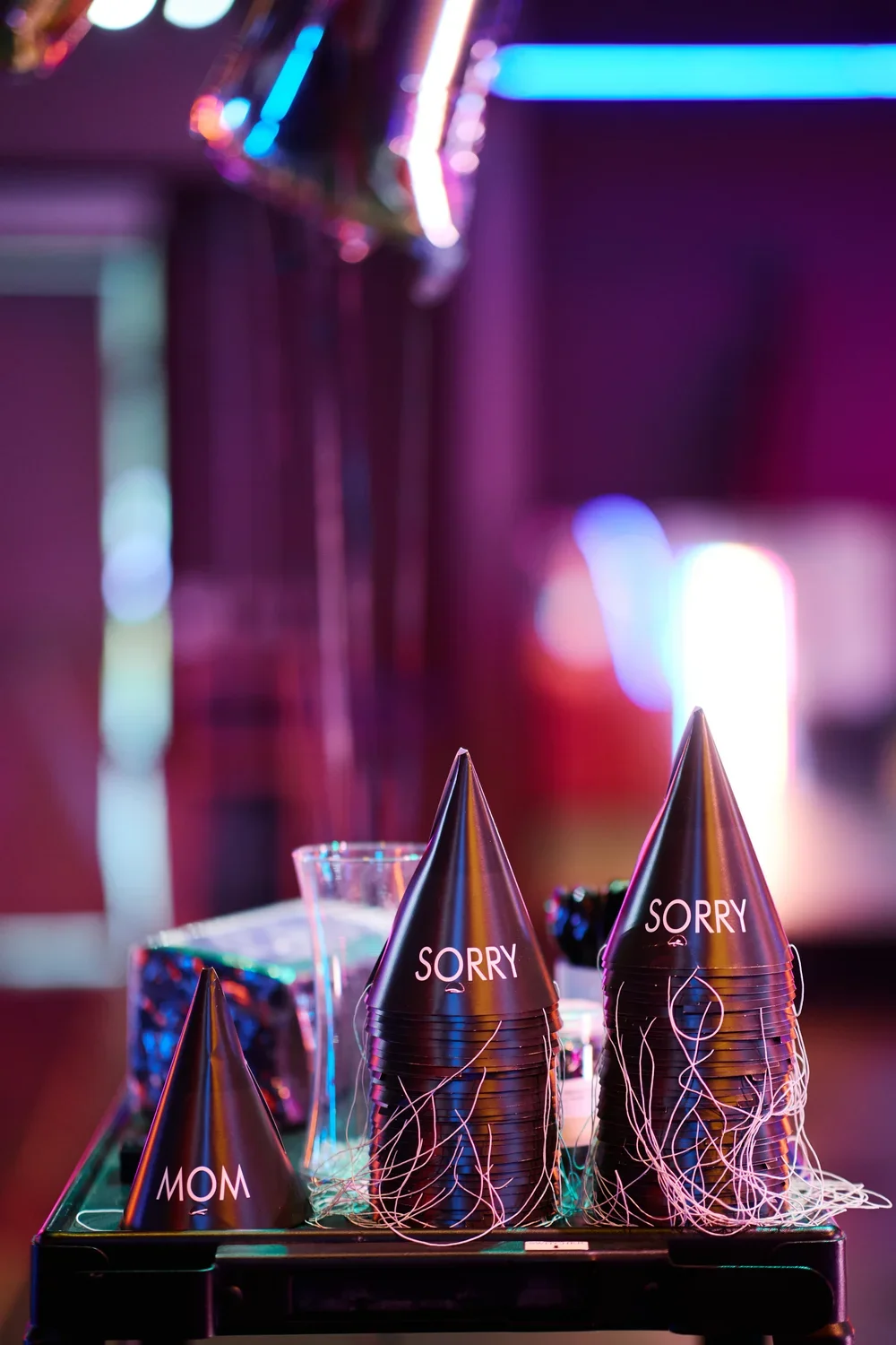 Black party hats are arranged on a dark surface. One hat says "MOM" in white letters, and the stacks of hats say "SORRY" in white letters. The background is blurred with purple and blue neon lights.