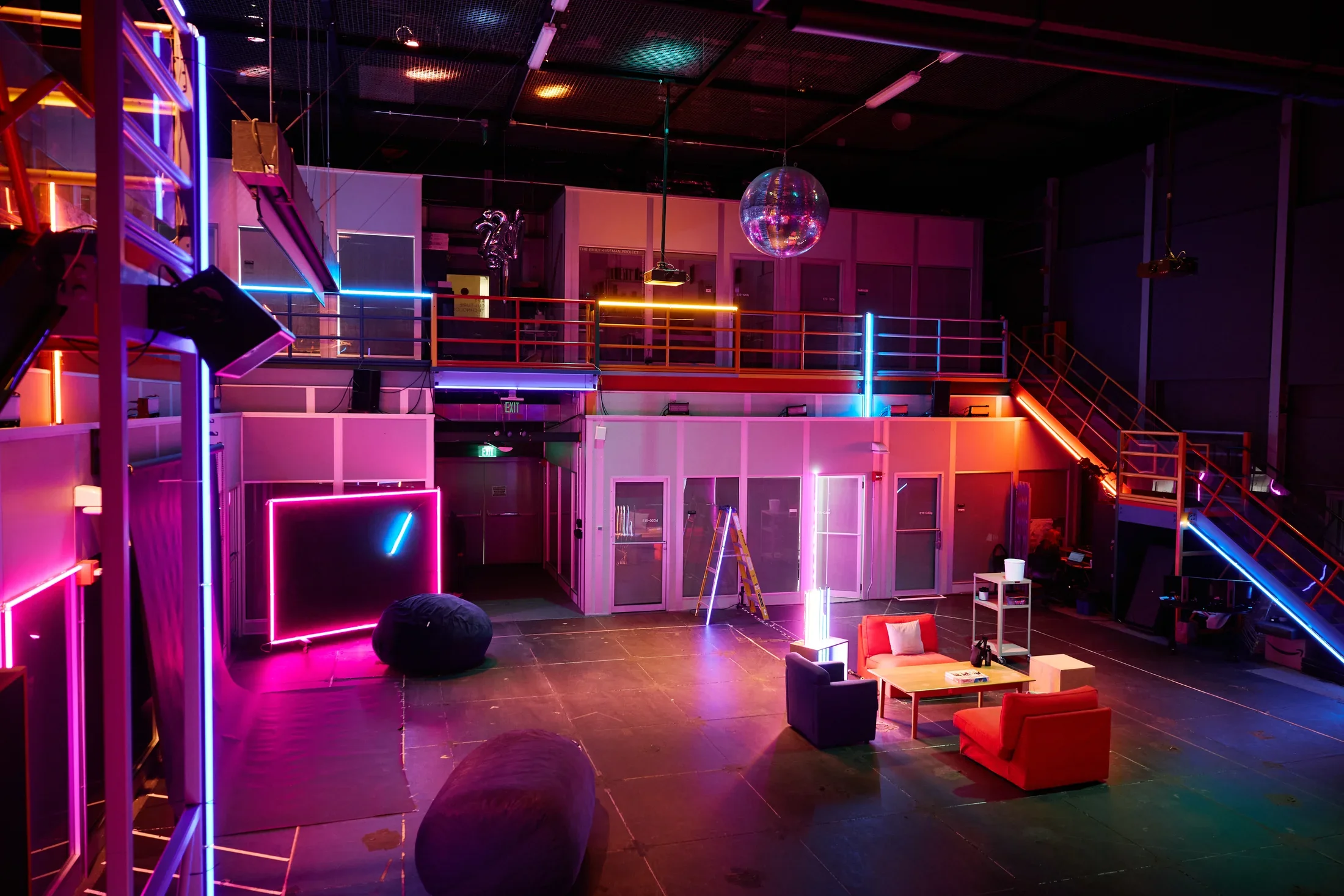 A large, dark space is transformed into a lounge-like performance space, illuminated by vibrant blue, pink, orange, and purple neon lights that trace the contours of the room, including a mezzanine and staircase. Furniture, including sofas, chairs, and bean bags, is arranged on the floor, with a disco ball hanging from the ceiling.