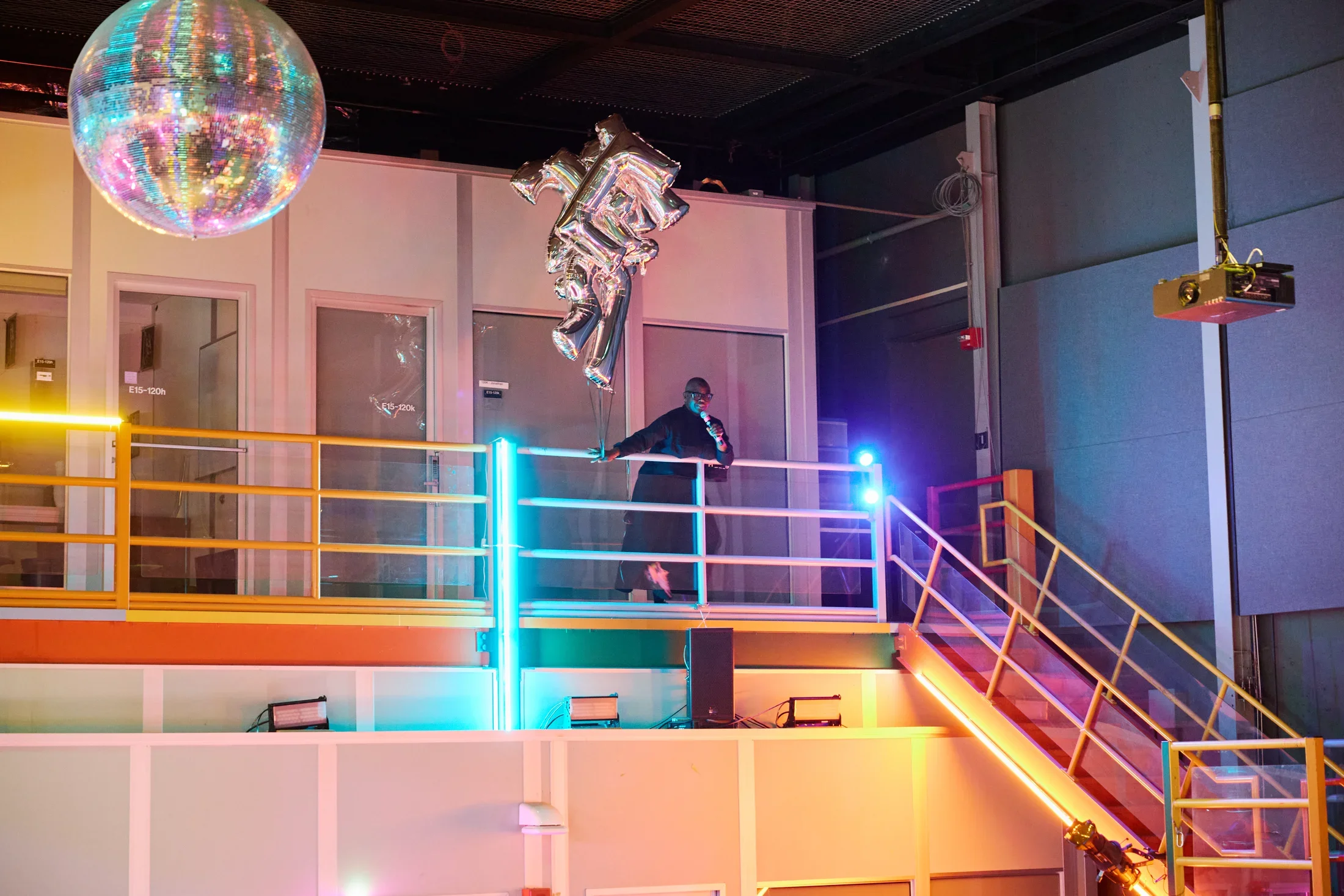A person in dark clothing stands on a metal mezzanine, holding a cluster of silver balloons shaped like large letters. A disco ball hangs from the ceiling, and the industrial-style room is illuminated by blue and orange tube lights.