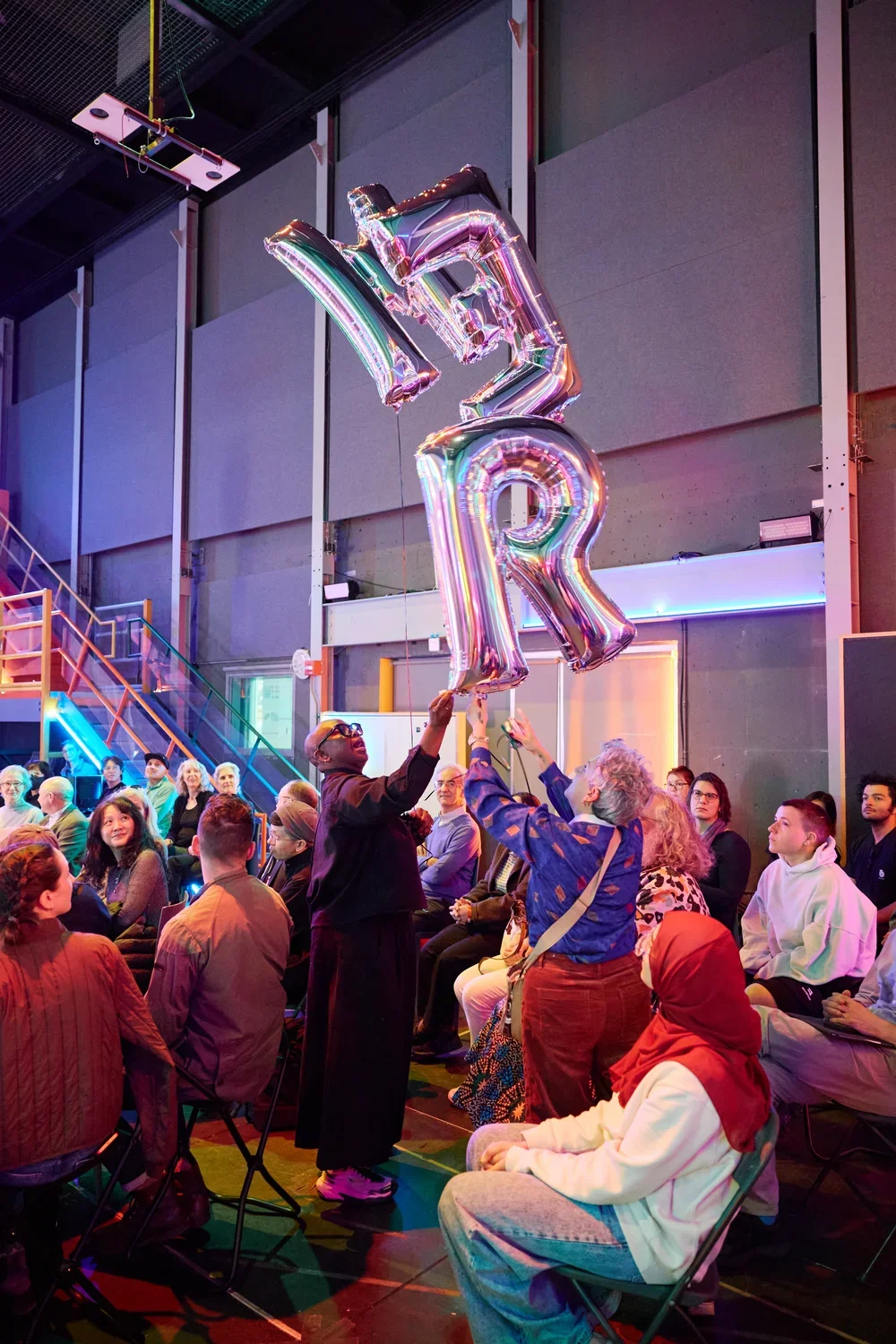 Two people hold up large, shiny, silver helium balloons (including the letters M, E, and R); perhaps one is handing the balloons to the other. An audience is seated around them, and colorful neon lights illuminate the background.