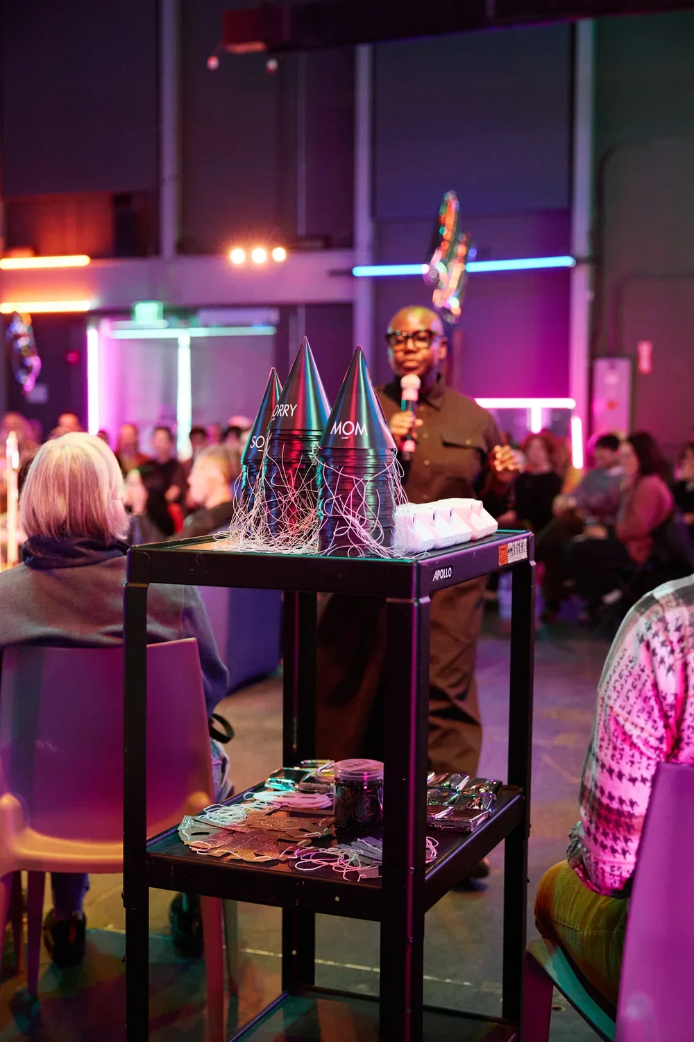 A person in dark clothing and glasses speaks into a microphone, standing behind a black tiered cart. On the cart's top shelf are three stacks of party hats that read "SORRY" and "MOM." An audience is seated in the colorful, neon-lit background.