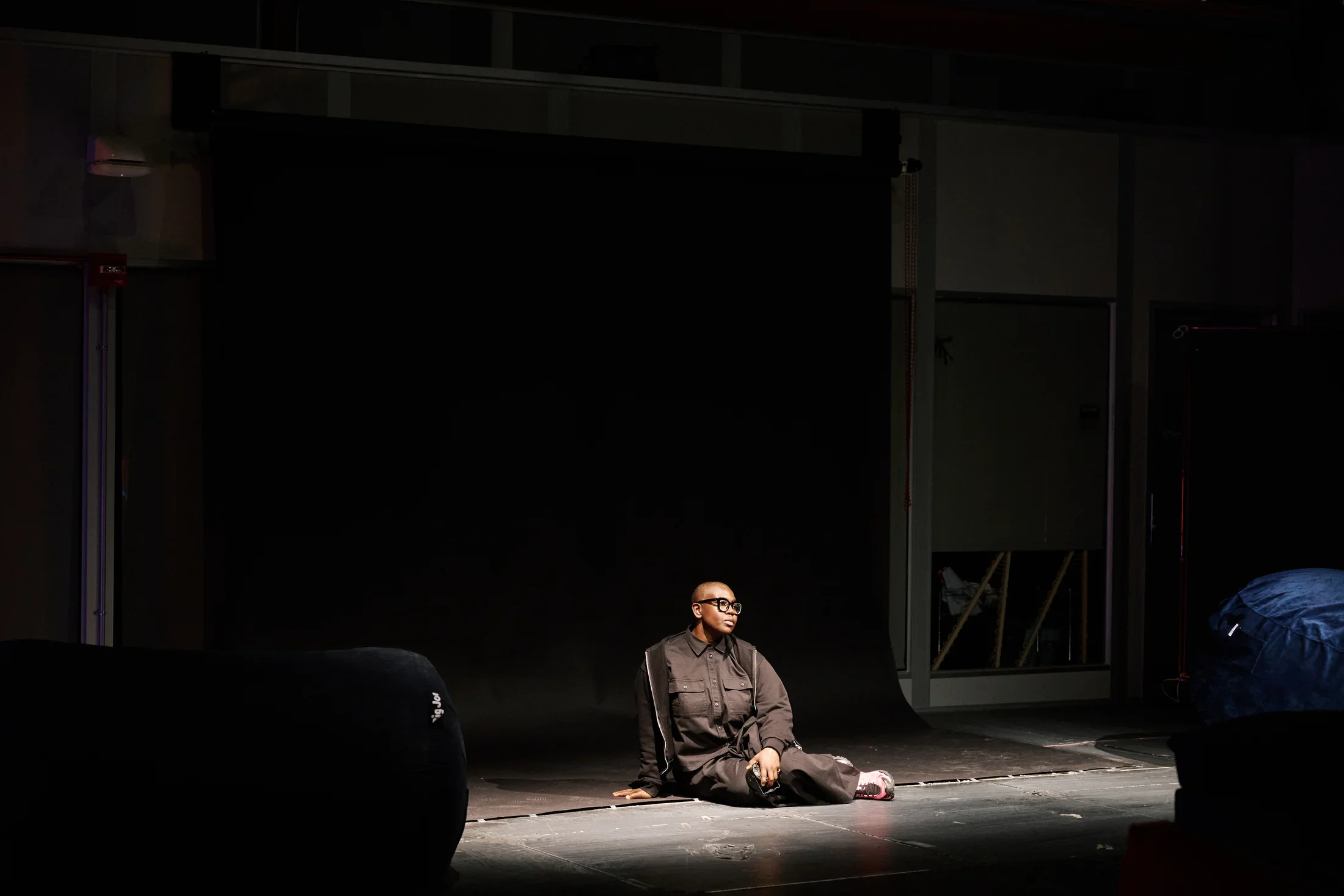 A person wearing black clothing and black glasses sits on a dark stage floor, looking to her left. She sits on a large black backdrop that extends from the wall to the floor, illuminated in a pool of light.