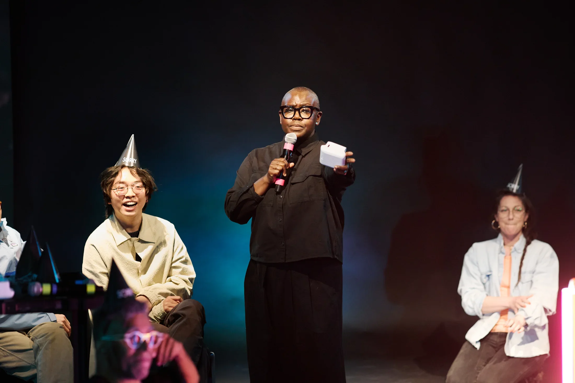 A person with glasses and a dark shirt stands center stage, speaking into a microphone and holding a white instant camera. She is flanked by smiling audience members, both wearing pointed black party hats. 