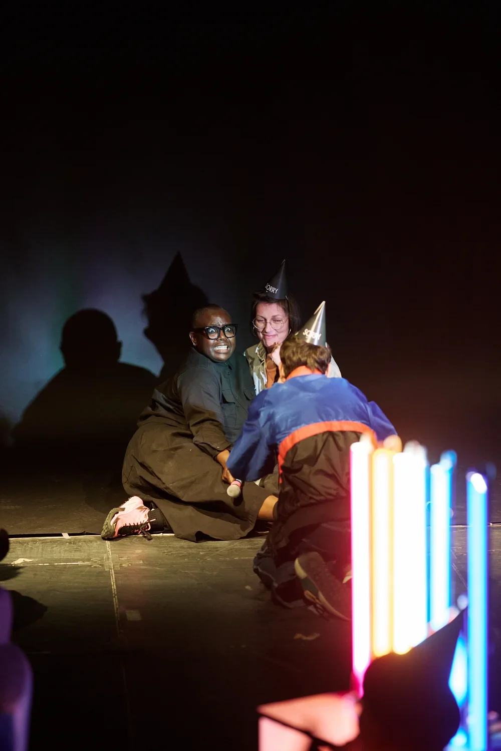 Against a dark backdrop, a performer dressed in black makes an exaggerated face—a smile stretched into a look of surprise or fright—while sitting next to a crouched audience member wearing a party hat. Another audience kneels in front of them, leaning forward to take a picture. Glowing vertical light tubes stand in the foreground on the right.