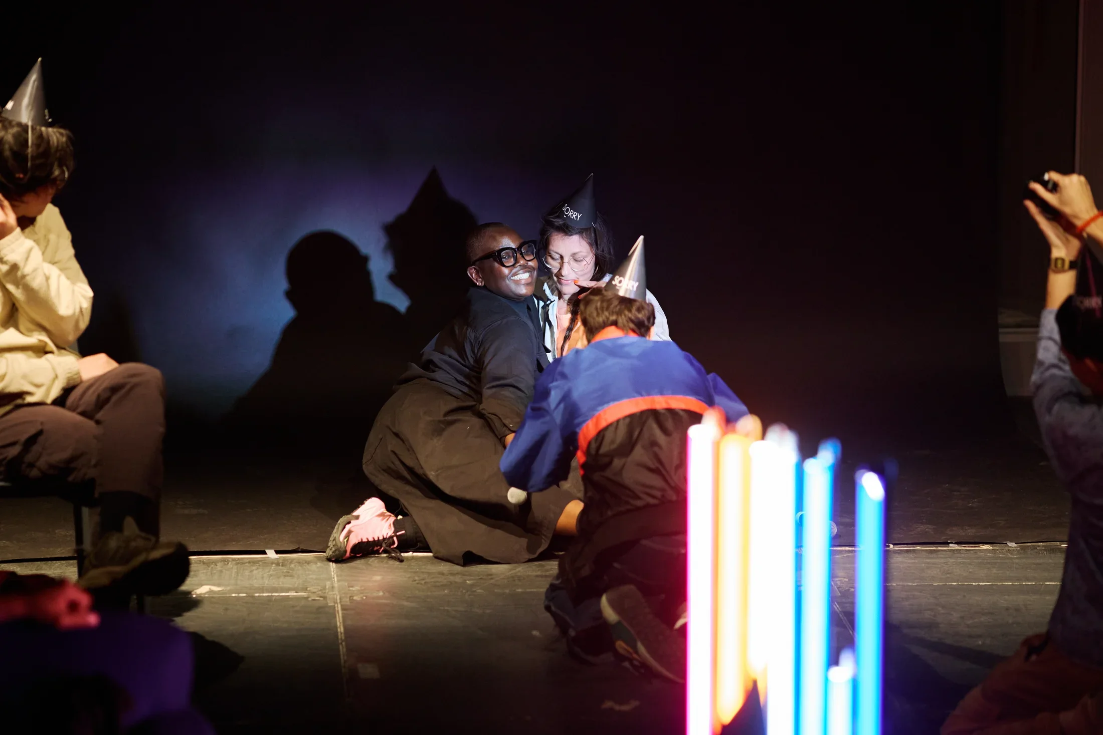 Two people are posed together onstage against a dark backdrop, one smiling broadly, the other looking downward. An audience member kneels in front of them, leaning forward to take a picture. Glowing vertical light tubes stand in the foreground on the right, with additional figures partially visible along the picture’s edges.