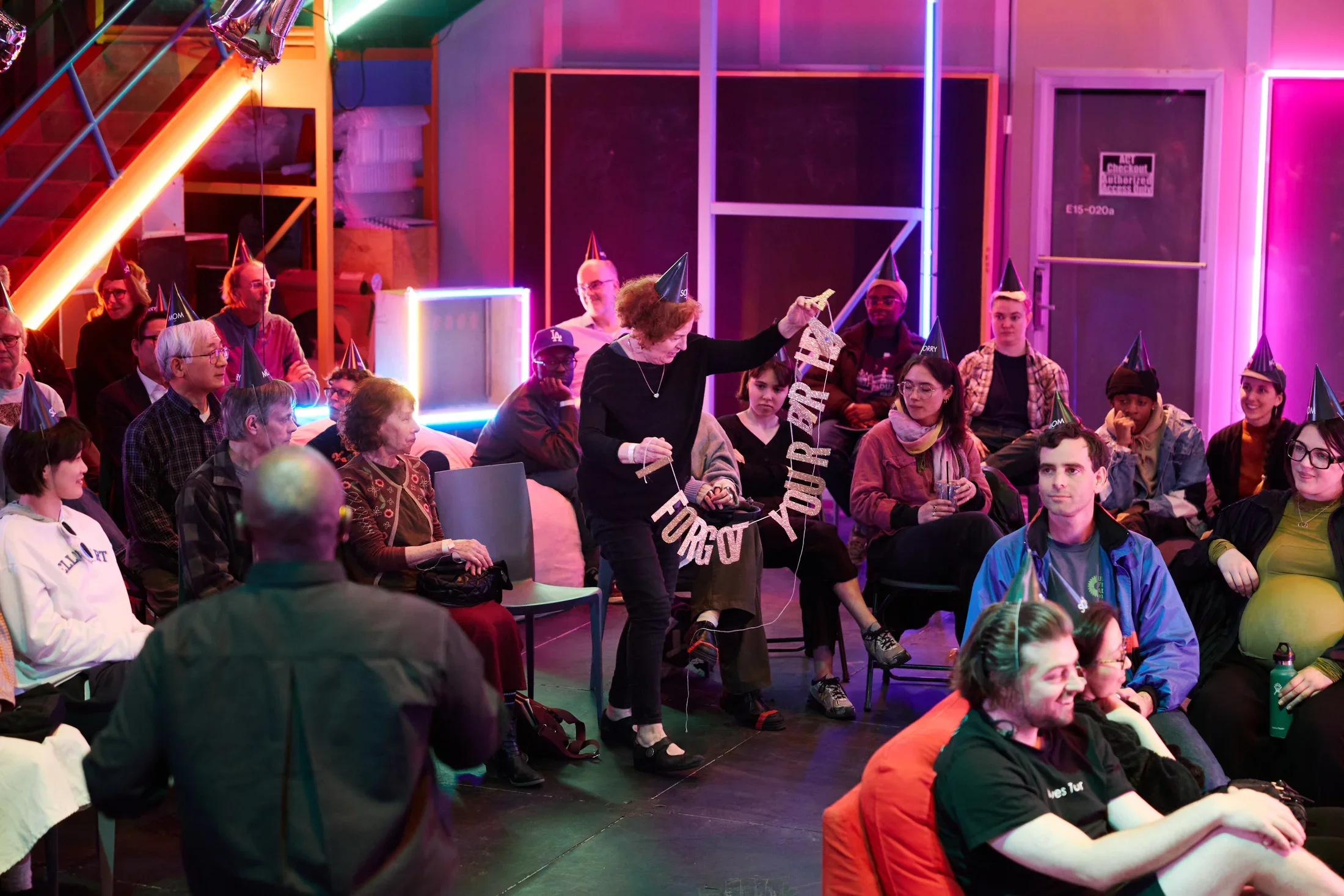 A group of people, many wearing dark party hats, are gathered in a room lit with pink and blue neon lights. A woman with red hair stands up from her seat holding a glittery silver banner of strung letters reading 'FORGOT YOUR BIRTHDAY.’ 