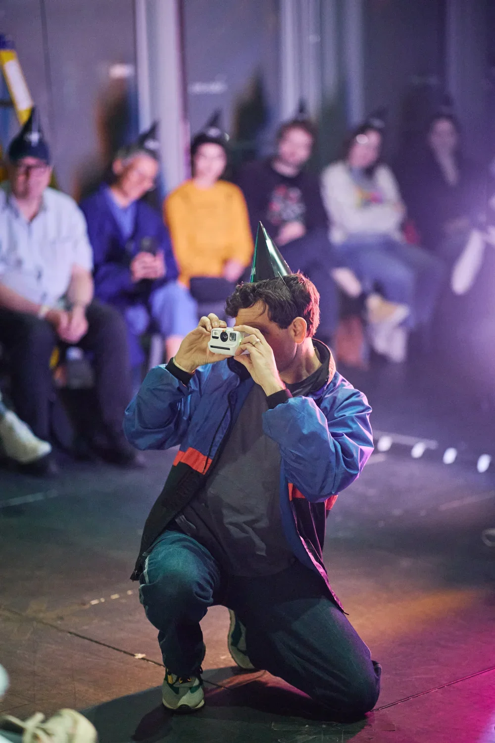 A man wearing a black party hat and a blue and black jacket kneels, holding a white instant camera to his face. Several people in party hats are seated in the blurred background.