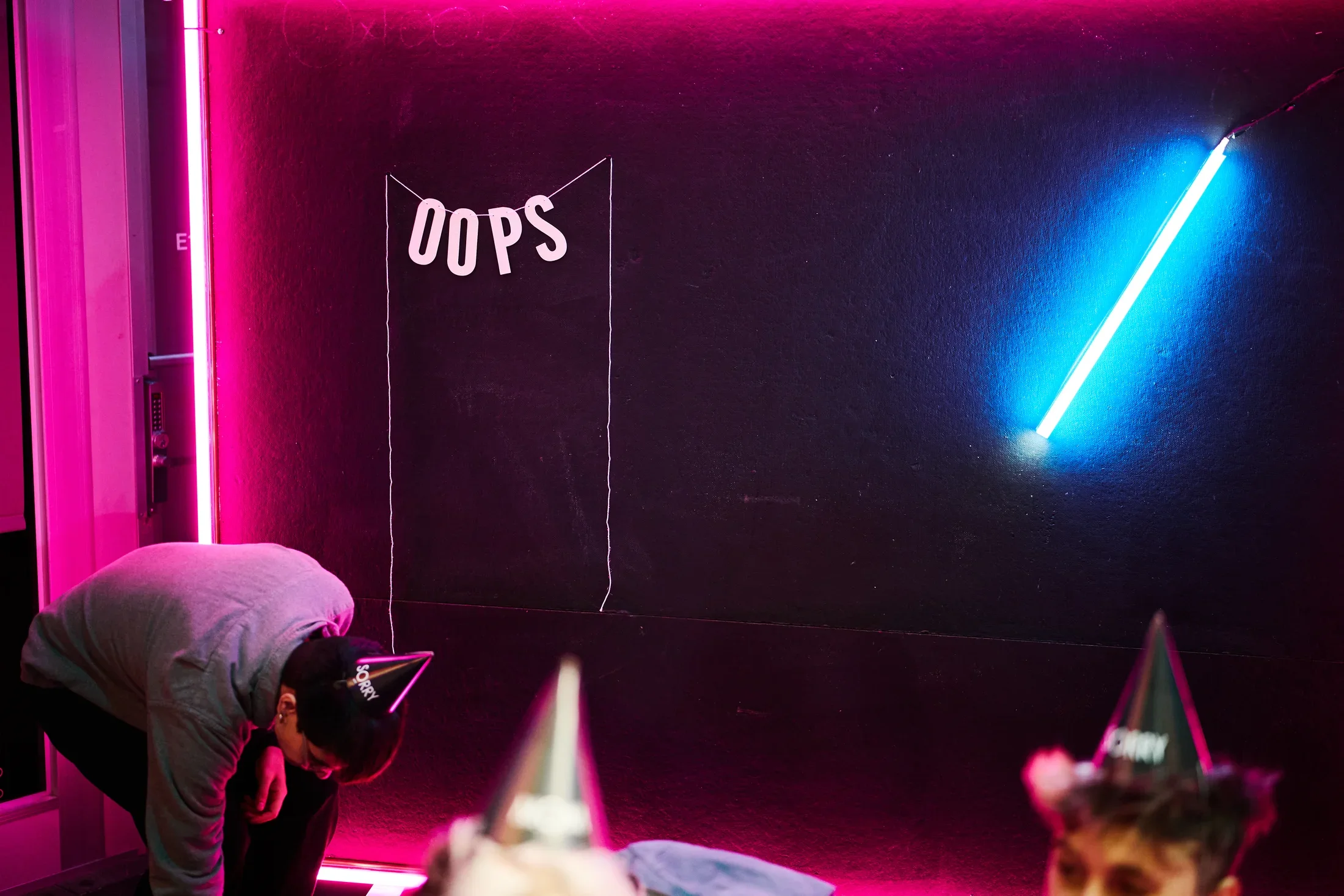 A dark room is illuminated by bright pink and blue neon lights. A banner with the letters "OOPS" strung together is hung on the wall, alongside a diagonal slash of blue tube lighting. Two people wearing pointed party hats are partially visible in the foreground.