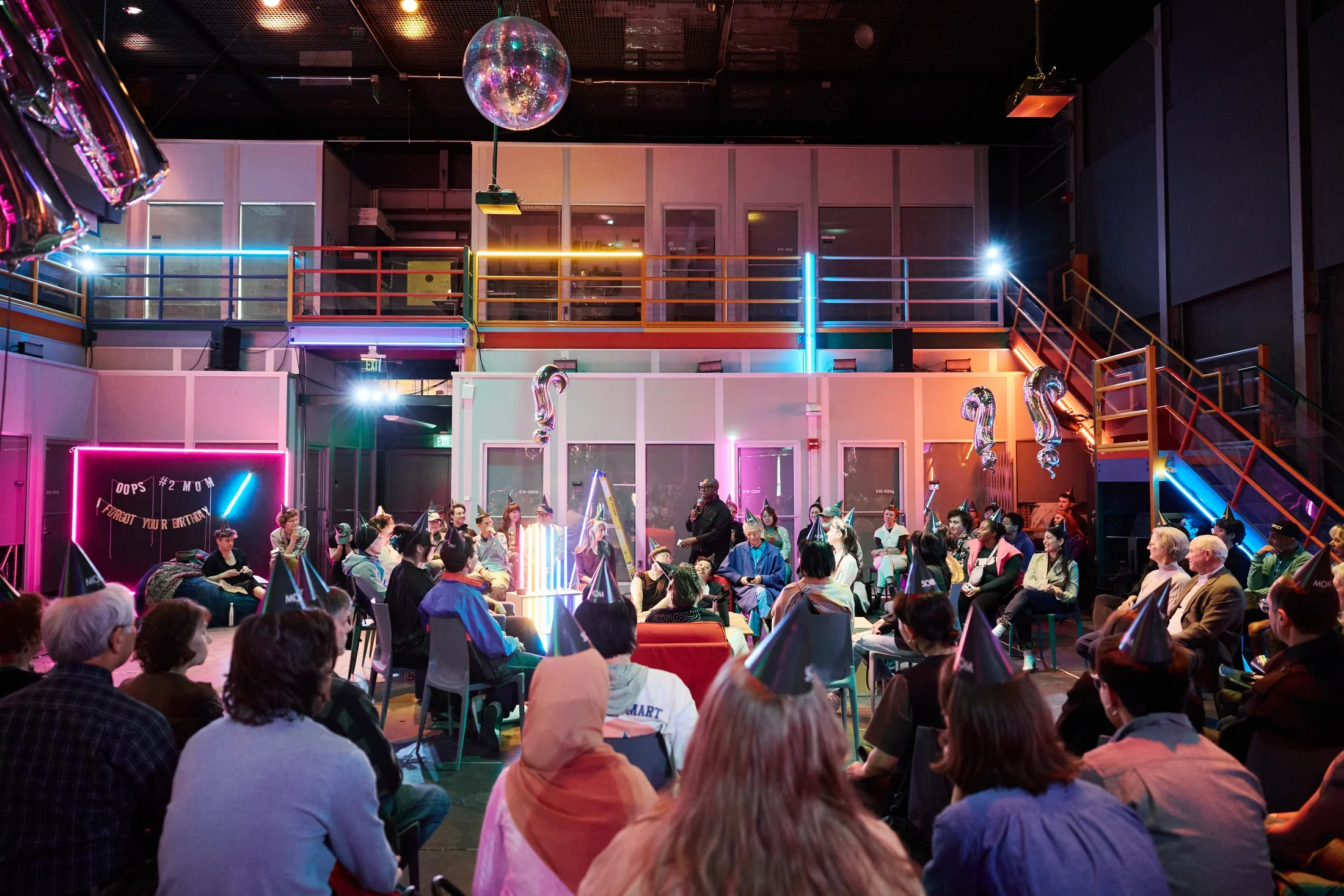 A large group of people, many wearing conical party hats, are gathered in a performance space. A speaker stands at the front addressing the seated audience, while neon lights, a disco ball, text banners, and silver balloons shaped like question marks decorate the multi-level room.