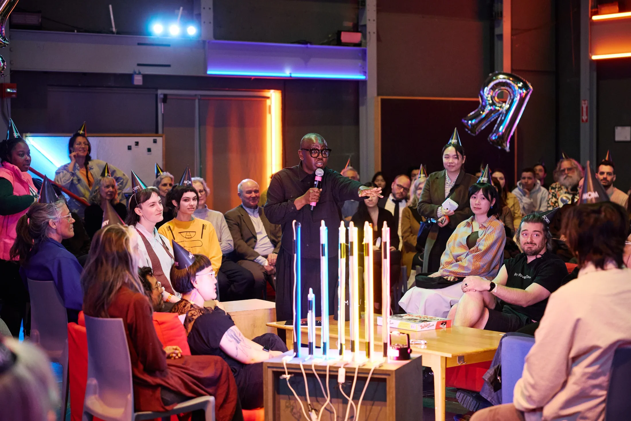 A performer in glasses and a dark shirt speaks into a microphone, gesturing to a seated audience. Many attendees wear conical party hats, and a large silver balloon shaped like the letter R visible on the right. Colorful vertical light tubes glow on a table in the foreground.