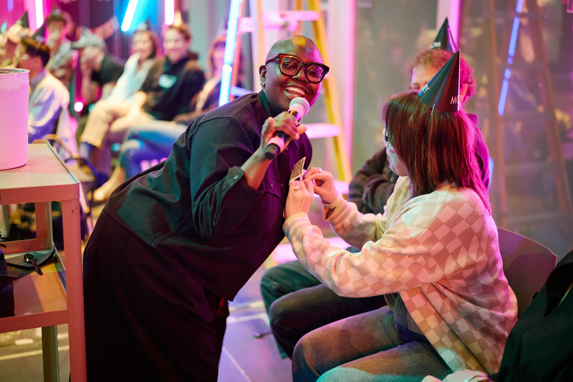 A person with short hair and dark glasses smiles while holding a microphone and leaning towards another person. The second person, wearing a party hat and a checkered sweater, pins a dollar bill to the performer’s shirt. Blurred figures and neon lights are visible in the background.