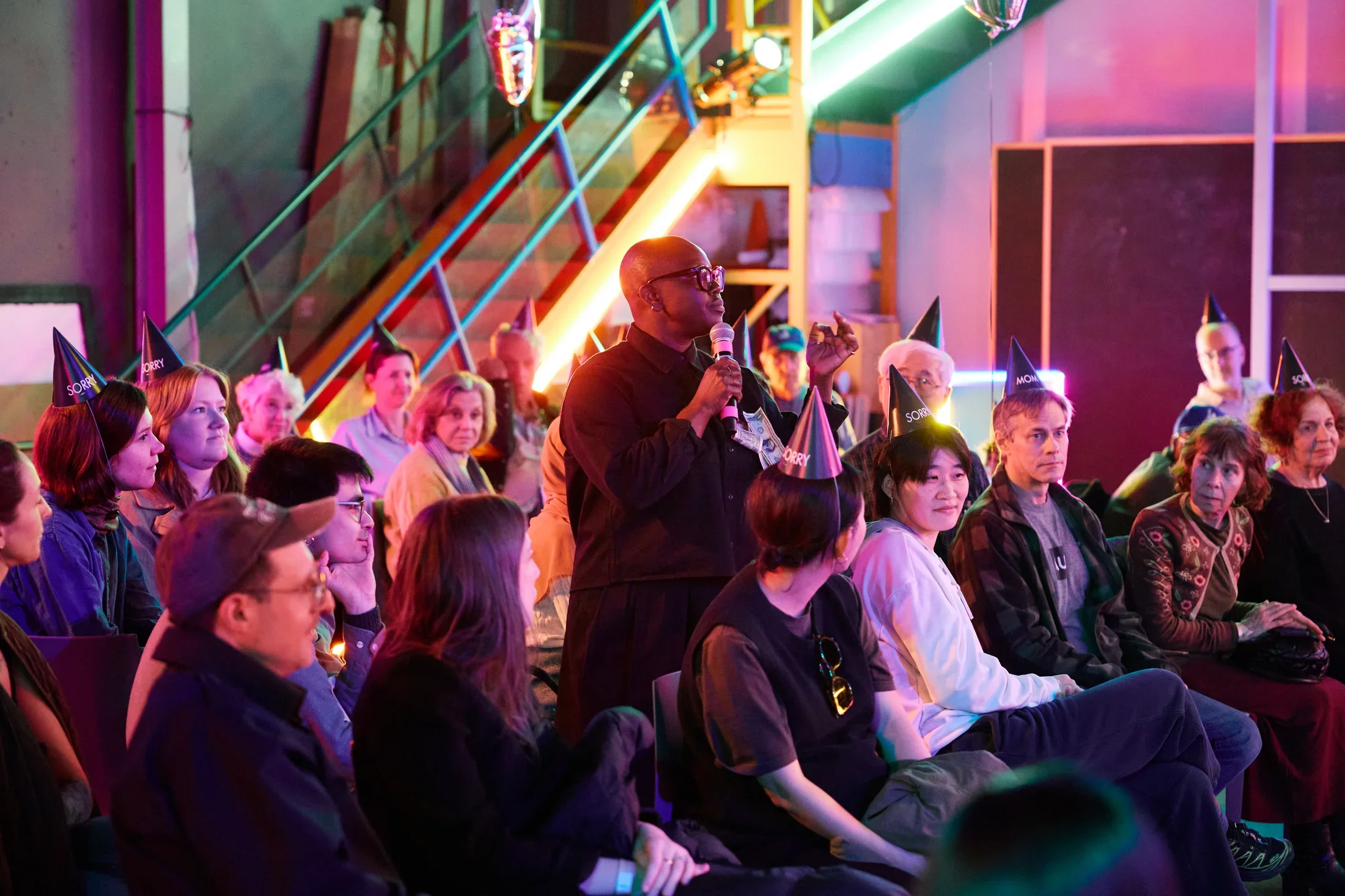 A person with a bald head and glasses, dressed in dark clothing, speaks into a microphone while standing in front of a seated audience. Many audience members wear black party hats with "SORRY" and “MOM” printed on them. The background features a metal staircase and neon lighting in pink and green hues.