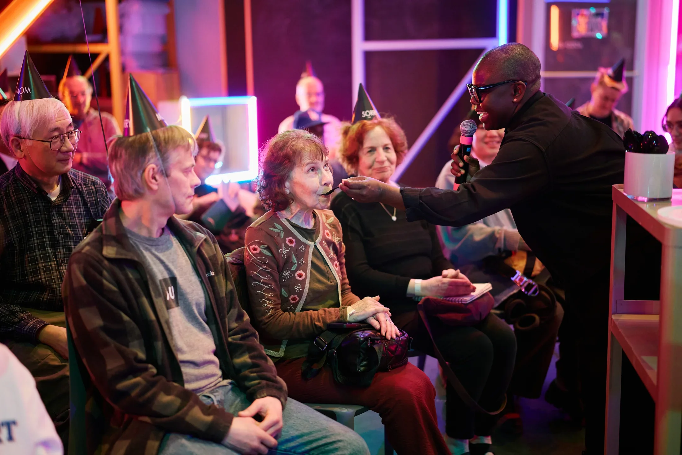 A person holding a microphone stands and feeds a spoonful of food to a seated older woman wearing a patterned cardigan. Several other people in black party hats are seated around them in an indoor setting with neon lights.