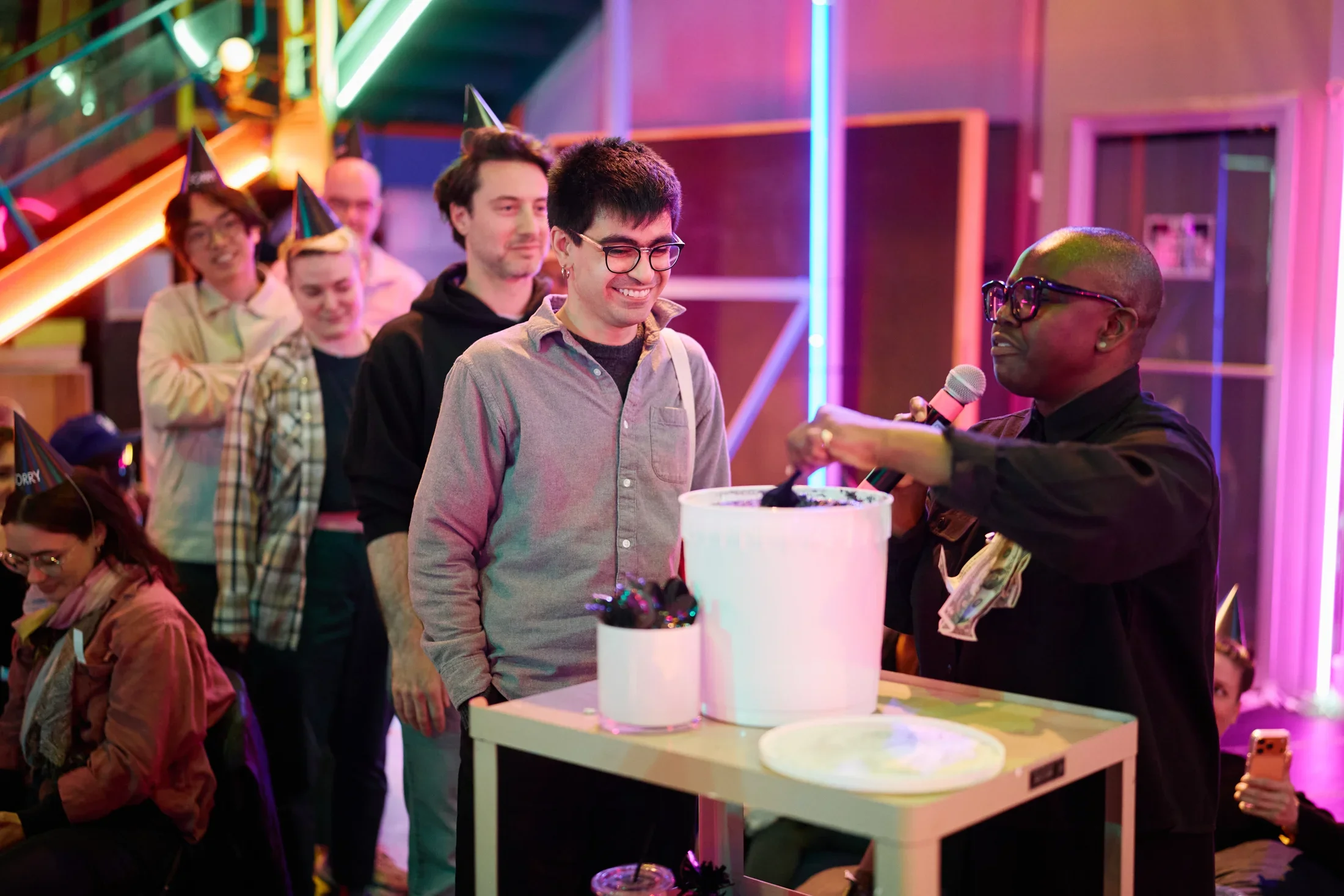 A performer wearing glasses with dollar bills pinned to her shirt scoops dark ice cream from a large white bucket. Several people, many wearing black party hats, stand in line to be served.