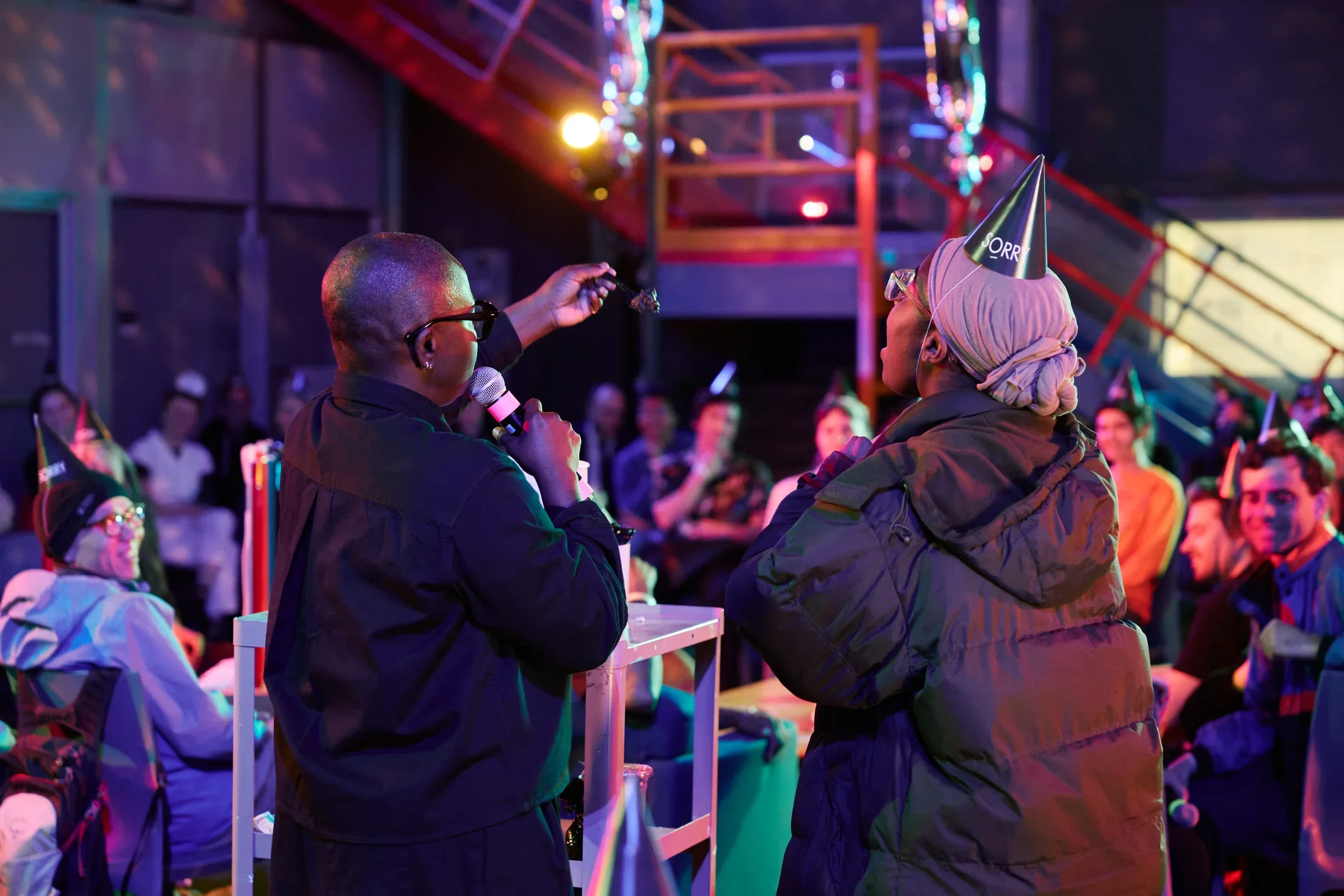 Two people face away from the camera towards a dimly lit room with colorful lighting and seated audience members. The person on the left, with a bald head and glasses, holds a microphone and extends a spoonful of dark ice cream towards the other person. The person on the right wears a party hat, a head covering, and a puffer jacket, leaning forward with an open mouth.