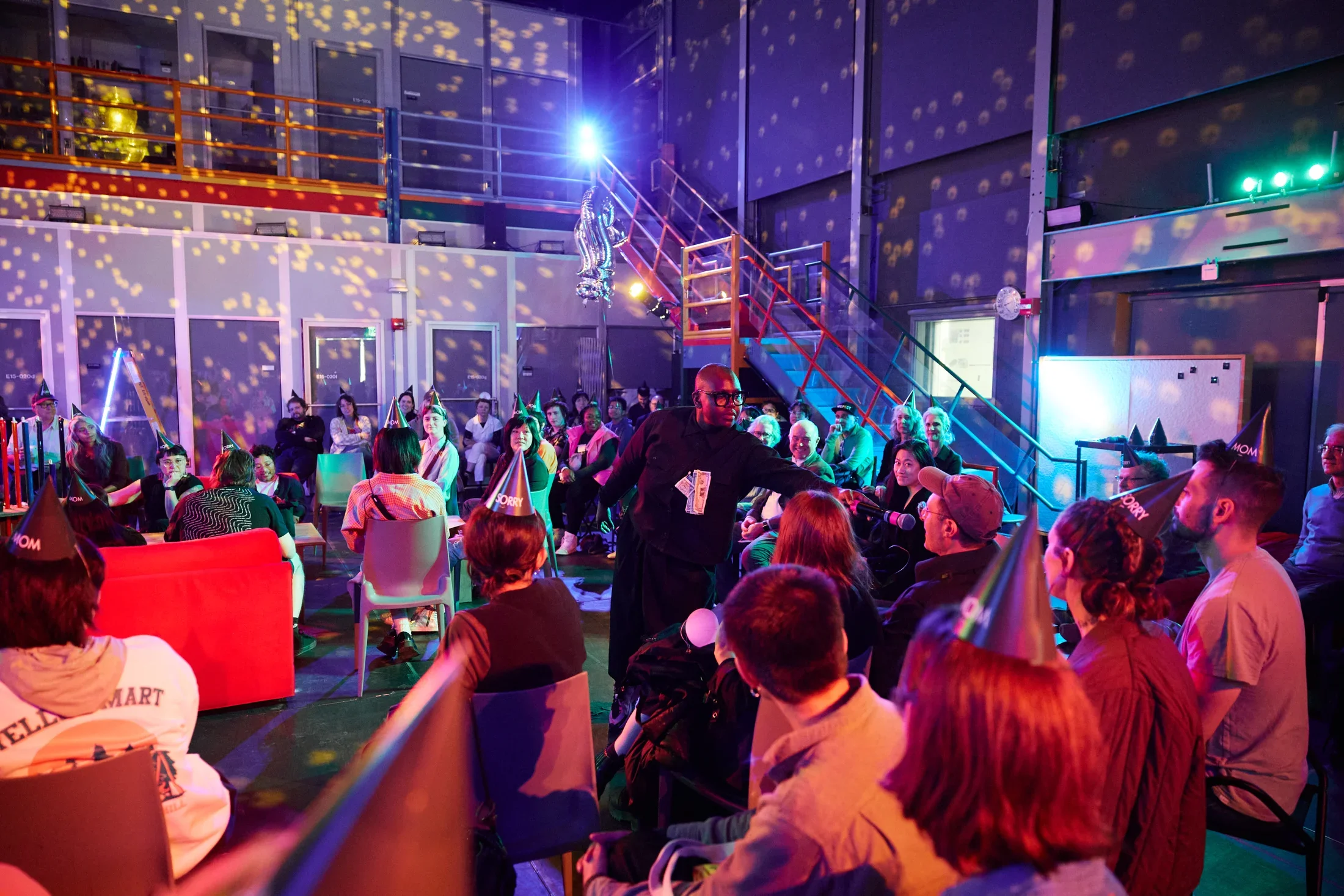 A person in a black outfit and glasses stands in the center of a performance space and holds a microphone to a seated man’s mouth. A few dollar bills are pinned to her chest. Many audience members wear pointed party hats, and the room is illuminated with multicolored lights and disco ball patterns.