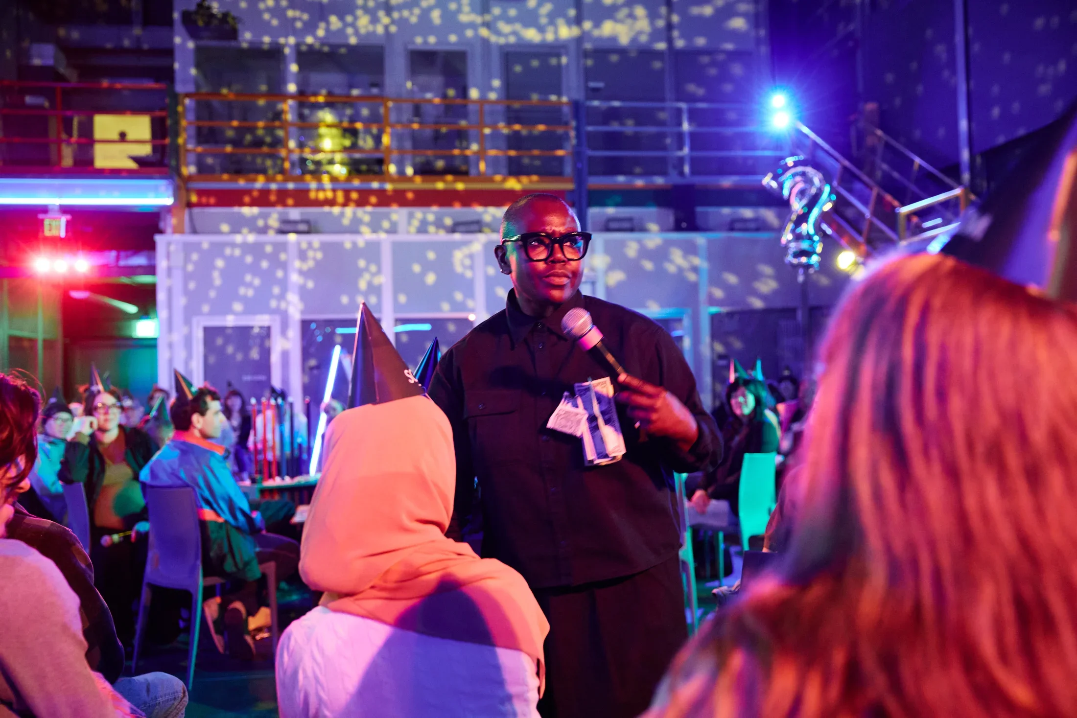 A person with dark-rimmed glasses and a black collared shirt speaks into a microphone. A few dollar bills are pinned to her chest. The room is illuminated by colored lighting and a disco ball, and audience members wearing party hats are visible in the background.
