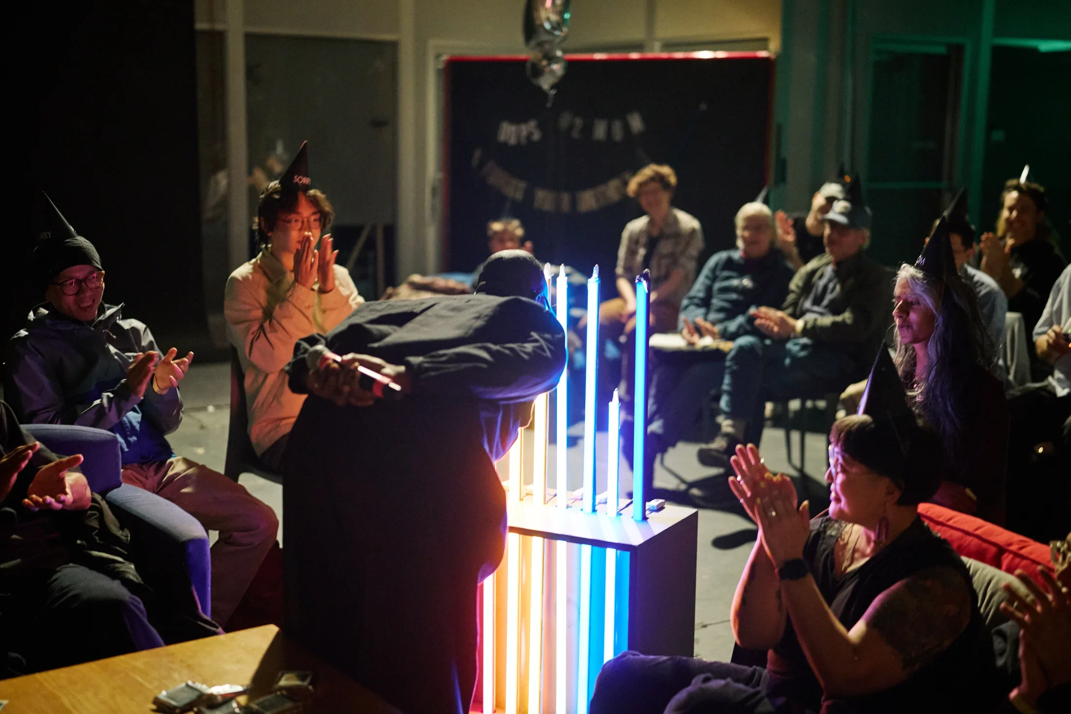 A group of people, many wearing pointed party hats, clap around a glowing light sculpture in a dimly lit room. The sculpture features vertical tubes emitting blue, purple, orange, and red light. A person in dark clothing leans over the illuminated sculpture, clutching a microphone behind her back.
