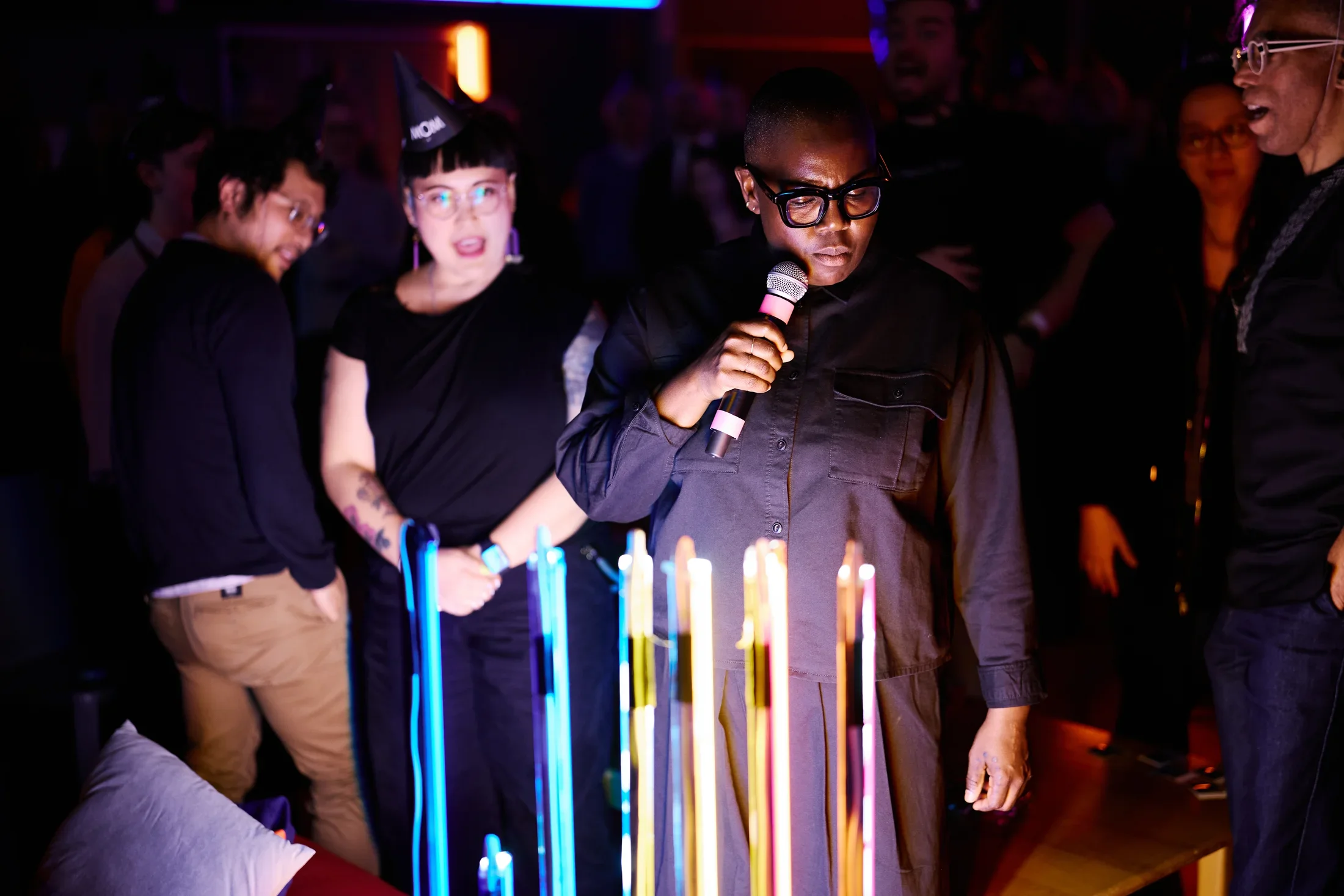 A person in dark clothing and glasses holds a microphone, speaking into it, while standing in front of several glowing vertical light tubes in blue, yellow, and pink. Other people are visible in the dimly lit background; one wears a party hat reading “MOM.”
