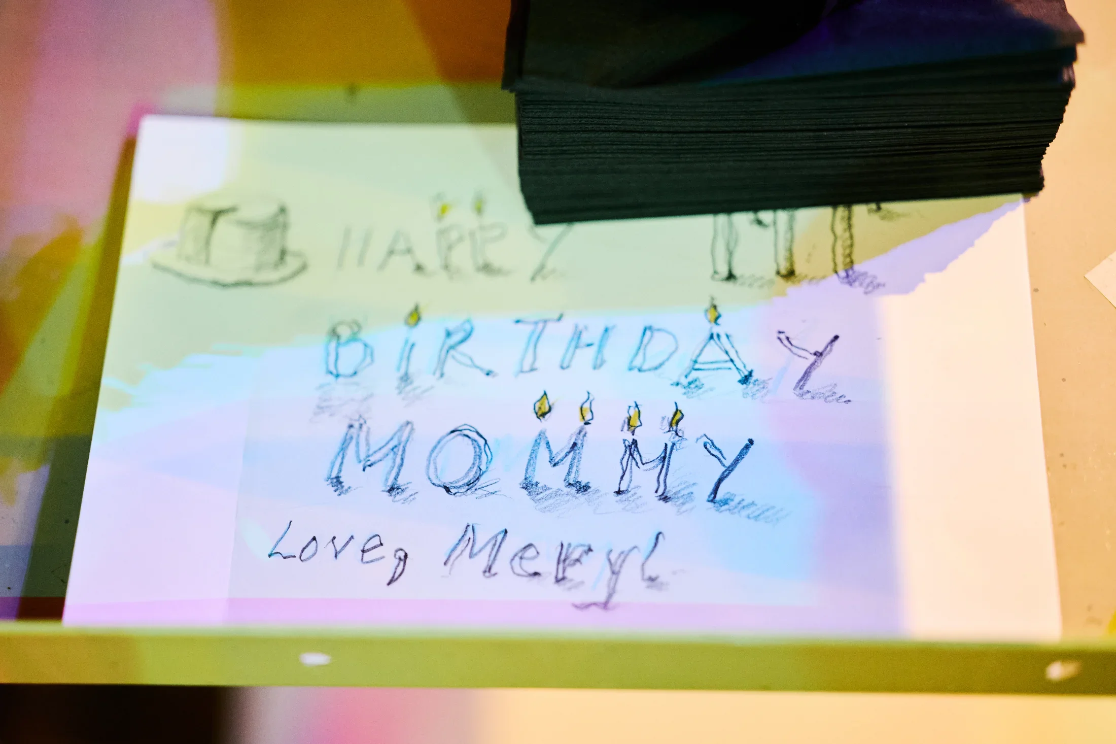A white sheet of paper displays a handwritten message in blue ink: 'HAPPY BIRTHDAY MOMMY Love, Meryl.' Small candle flames are drawn above some letters, and a simple cake drawing appears near the top. The paper partially obscured by a stack of dark napkins in the upper right.