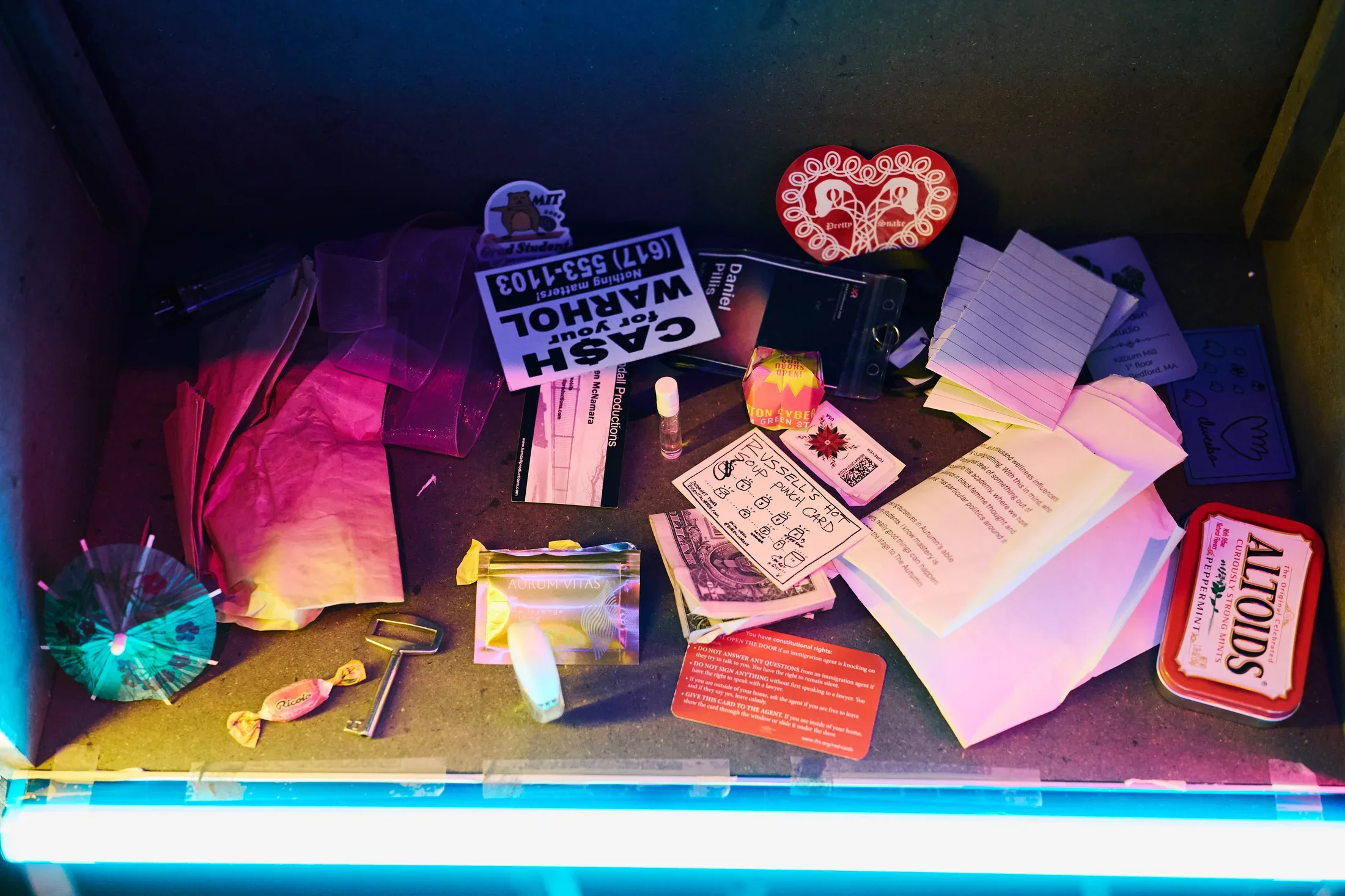 A collection of small items, including a green drink umbrella, a key, a dollar bill, a "CA$H for your WARHOL" sticker, crumpled pieces of paper, and an Altoids tin, are scattered on a cardboard surface
