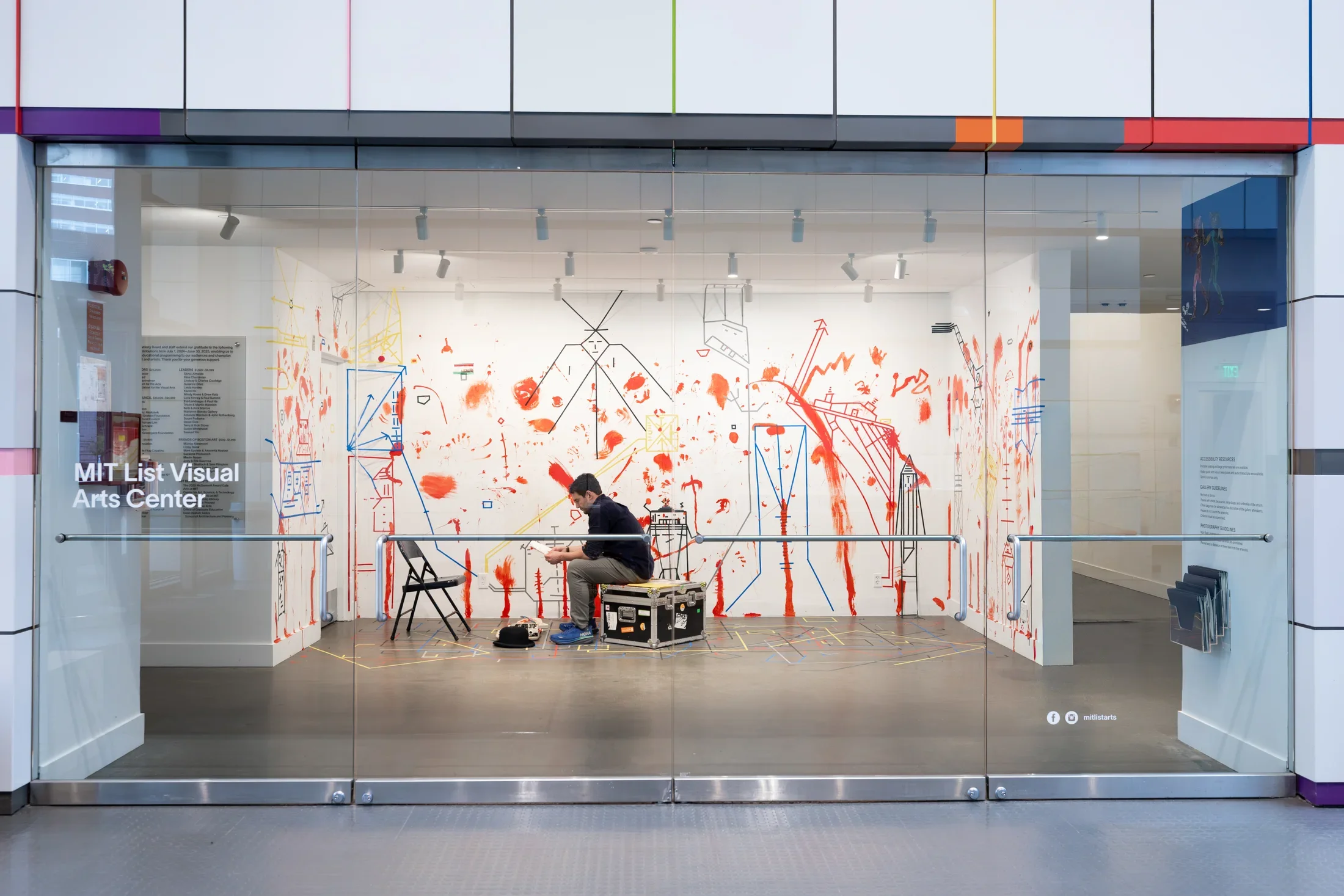A man sits on a sturdy chest or box and looks down at the packet of paper in his hands. The white walls behind him are marked with dense lines of colored tape and splotches and smears of red paint. The space is viewed through four glass doors; the far left one reads, “MIT List Visual Arts Center.”