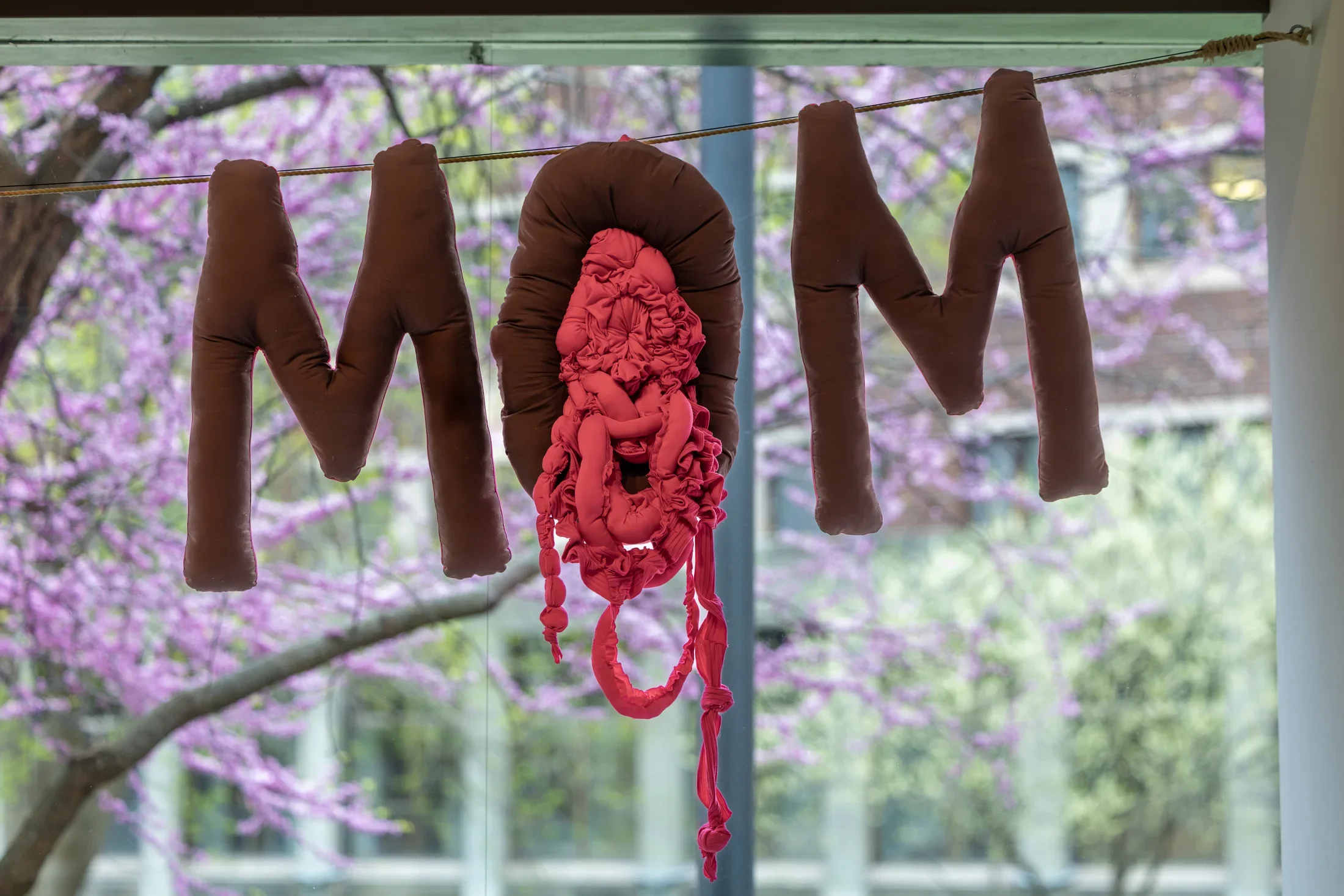 Three stuffed fabric letters spell 'MOM' and hang on a cable in front of a large window. The plush letters are brown or burgundy, and the middle of the ‘O’ is filled with a tangled mass of reddish-pink fabric that hangs down to reach the bottom of the frame.