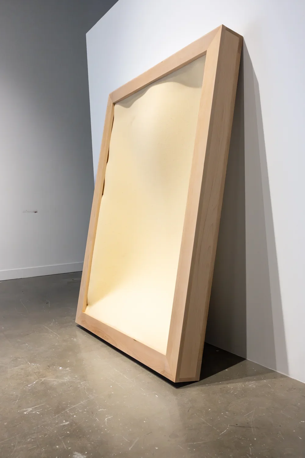 An open rectilinear wood frame encasing soft yellow foam stands vertically and leans against a wall. It is shown from the side at an angle, emphasizing the gap created by its lean.