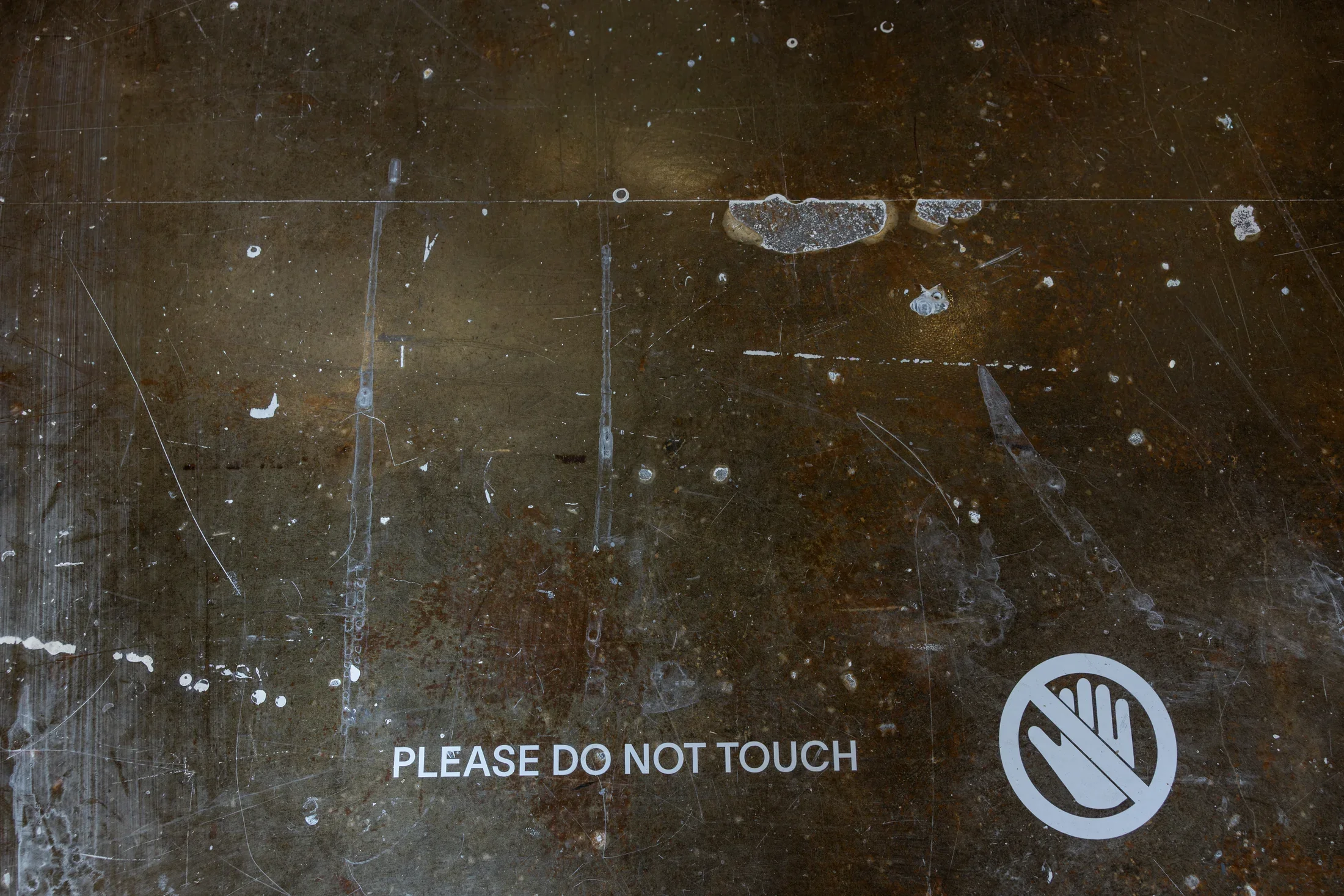 A dark floor, marked with scratches and drips of white paint, displays the white text 'PLEASE DO NOT TOUCH' next to a white circular symbol of a hand with a diagonal line through it.