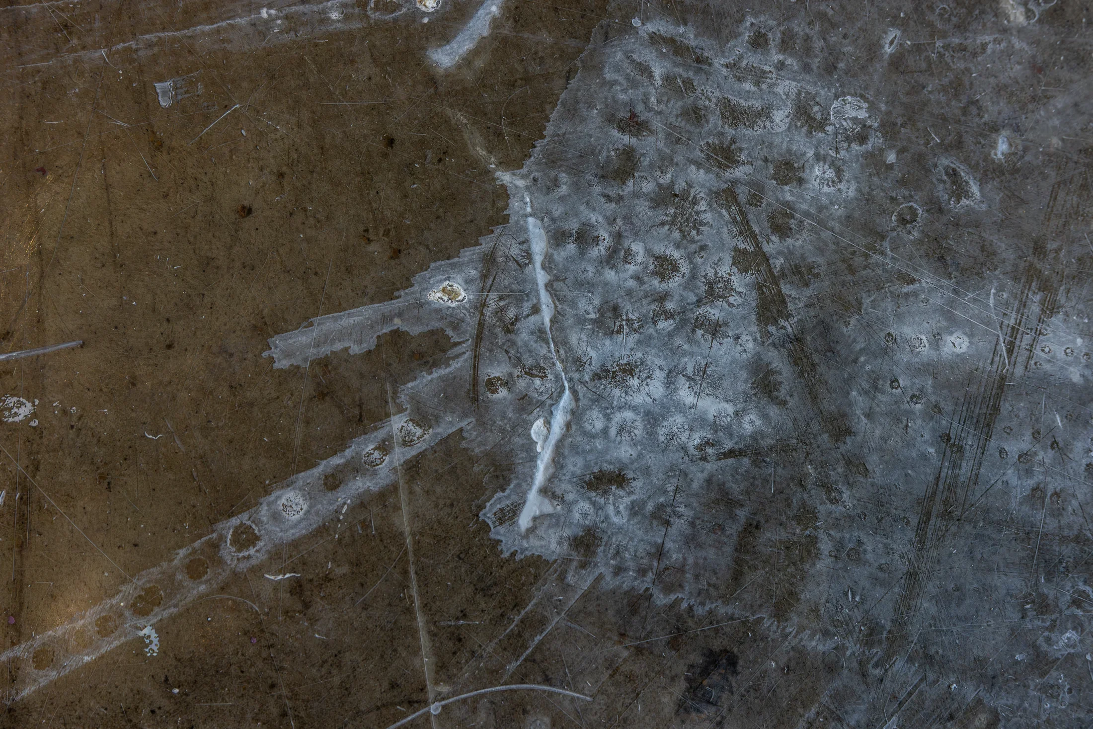 A dark, textured floor with white and gray residue, streaks, and numerous fine scratches. The surface has an uneven, mottled appearance.