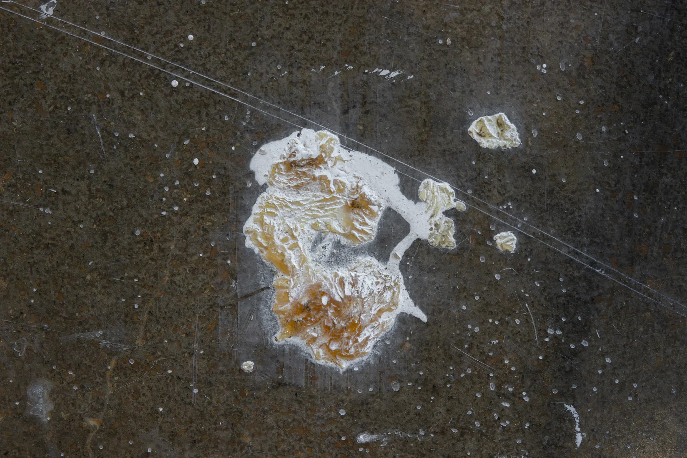 An irregular, textured mass of white and light brown material appears congealed on a dark, scratched floor. The dark background has numerous small white specks and thin linear marks.