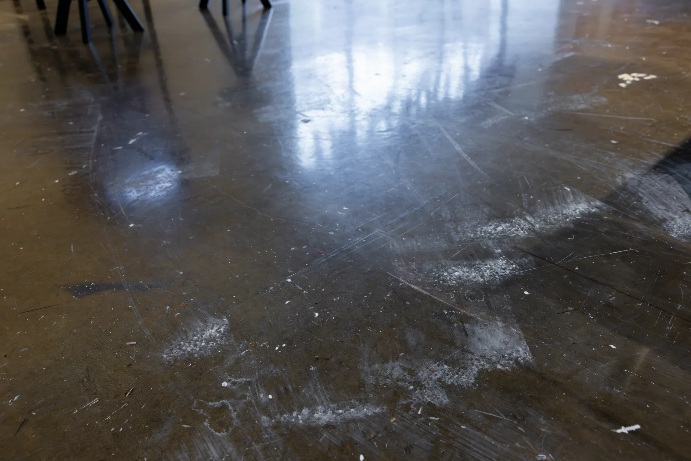 A dark, reflective floor is covered with numerous scuff marks and scattered white, drippy residue. Reflections of a window and the spindly legs of stools are visible towards the top of the frame.