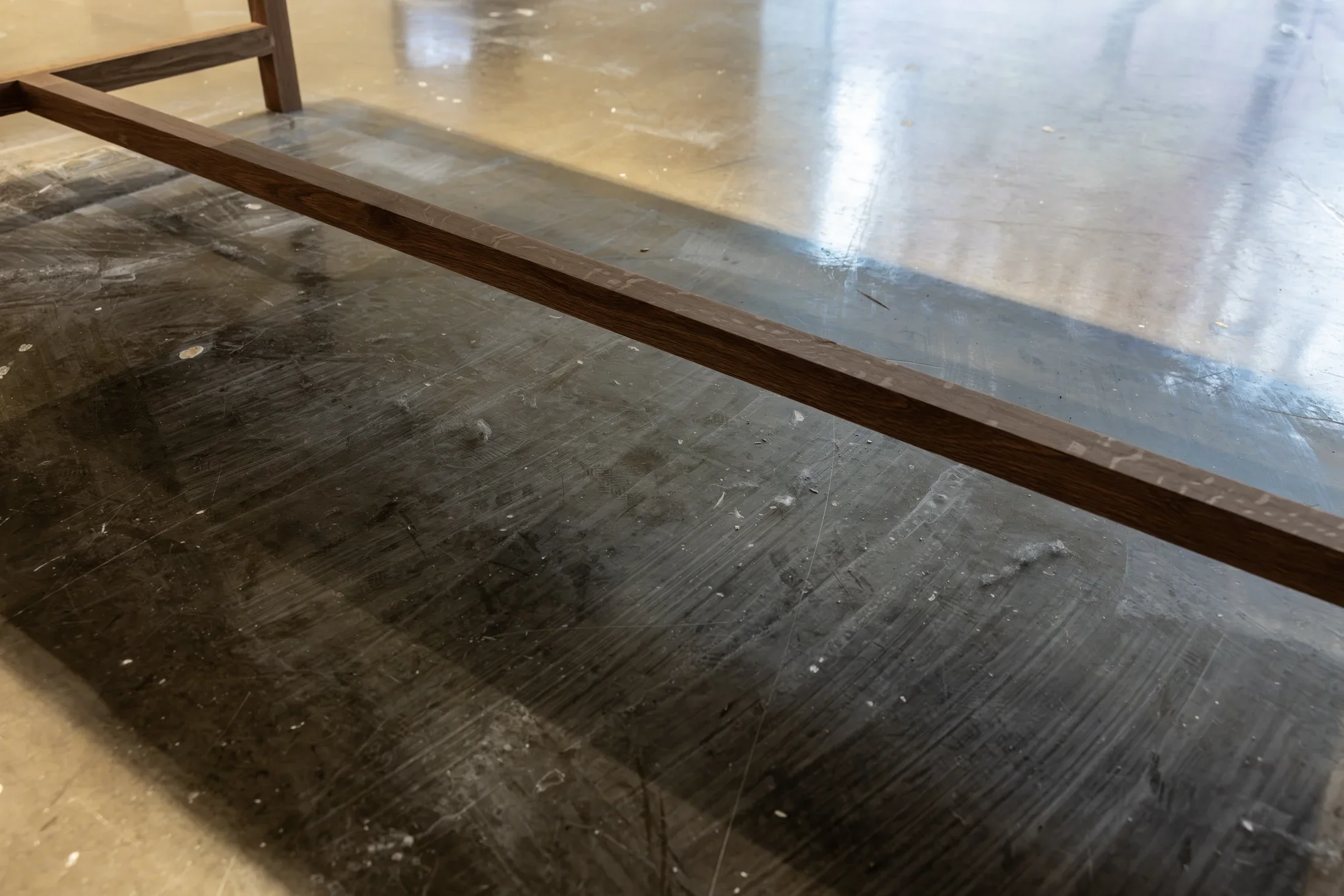 A dark wooden beam, the bottom of a freestanding vitrine, extends diagonally across a floor. Light striations, specks of dirt, and numerous dust bunnies sit on the floor underneath the vitrine.