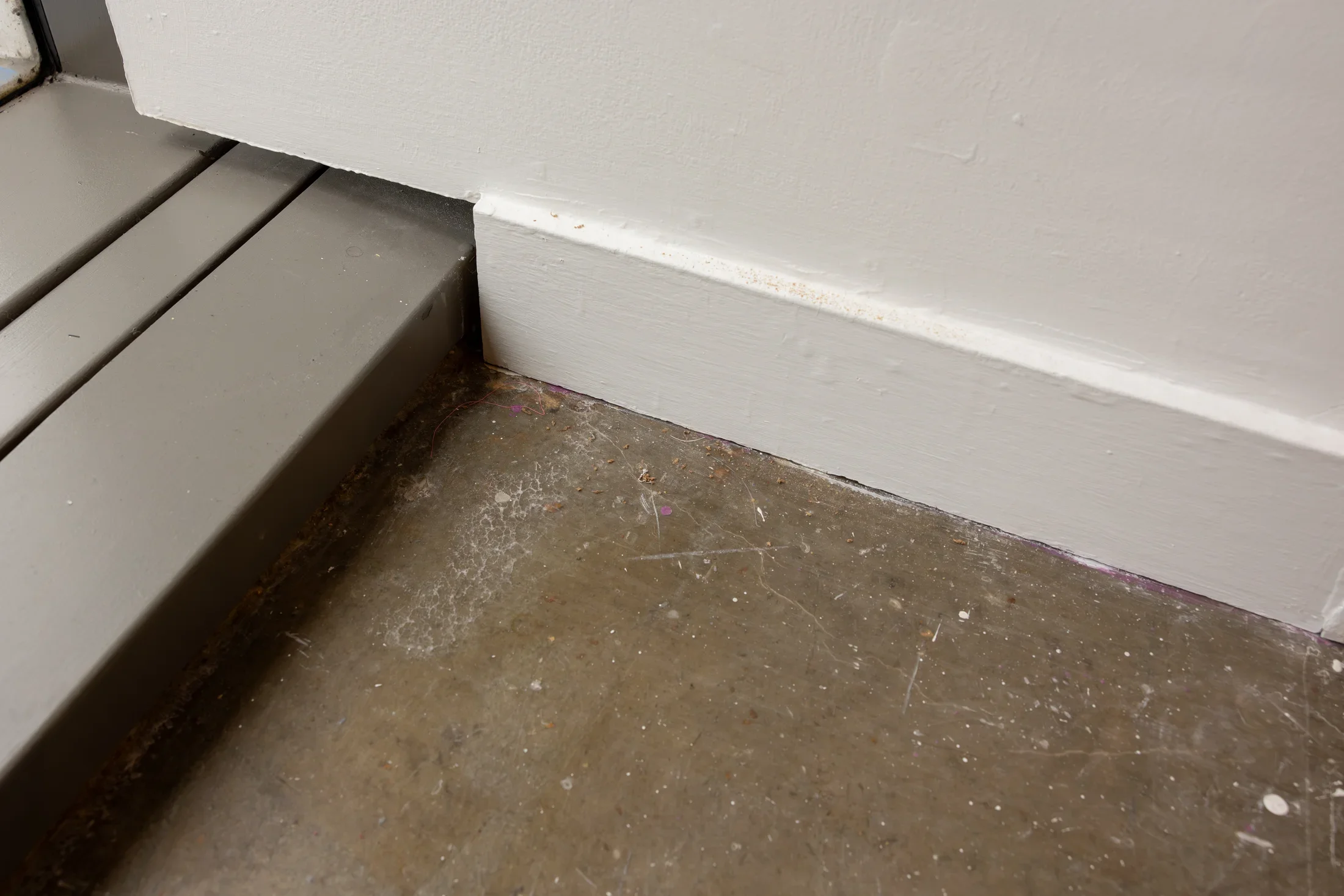 A close-up of a corner where a window frame meets a white wall and a gray floor. The corner is speckled with various light marks and dust.