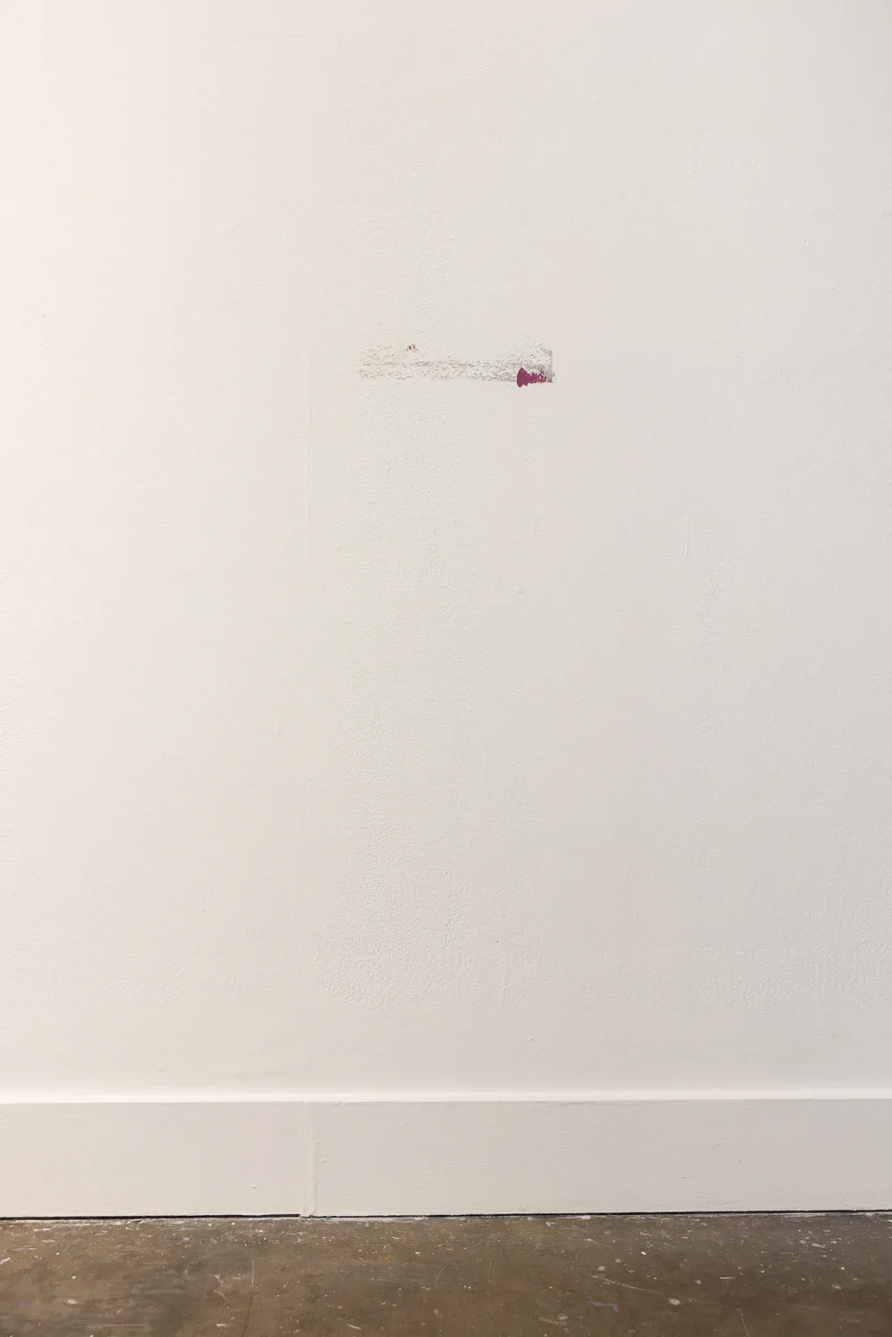 A white wall features a small, horizontal gray residue mark with a magenta paint splash on its right end. A white baseboard runs along the bottom, above a dark, mottled floor.
