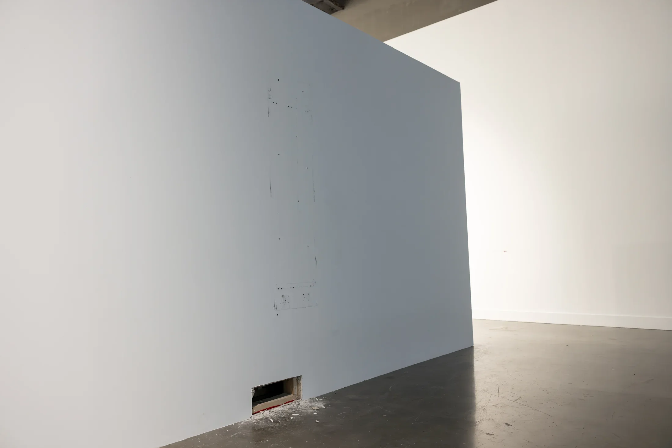 Scuffs and small holes outline an oblong, vertical, rectangular shape on an otherwise bare white wall. Near the floor, a smaller rectangle is cut from the wall entirely, spewing sawdust and scraps of wood onto the floor nearby. A second wall is visible behind the freestanding one.