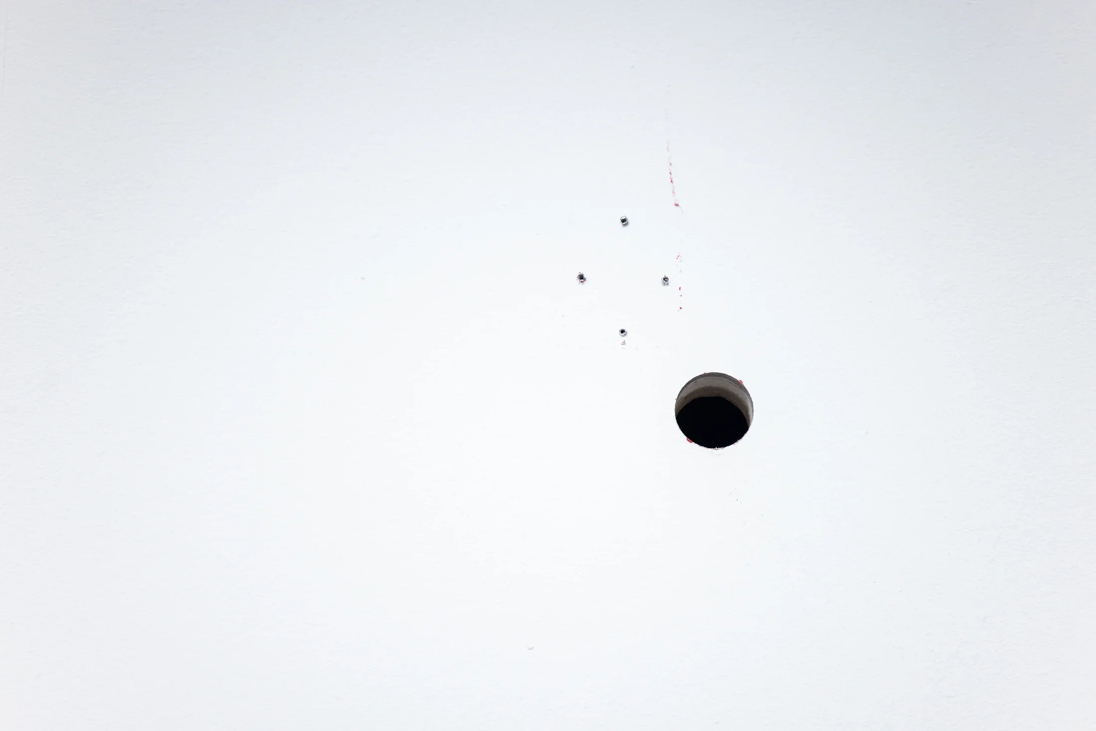 A white wall is punctured by a dark circular hole. Above the hole, four much smaller holes form a diamond shape.