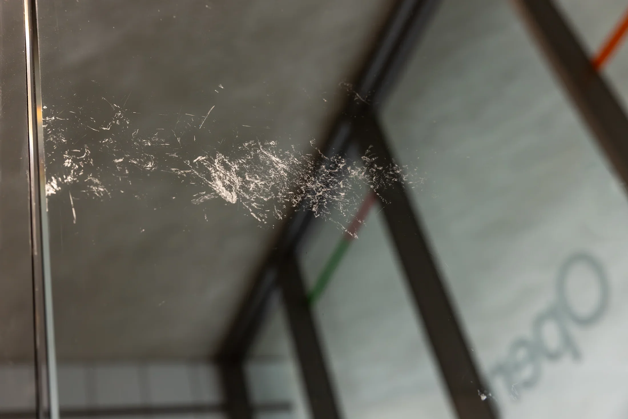 A close-up view of a glass surface covered in numerous fine, white scratches or marks. The blurry background shows an interior space with a concrete ceiling and dark architectural frames.