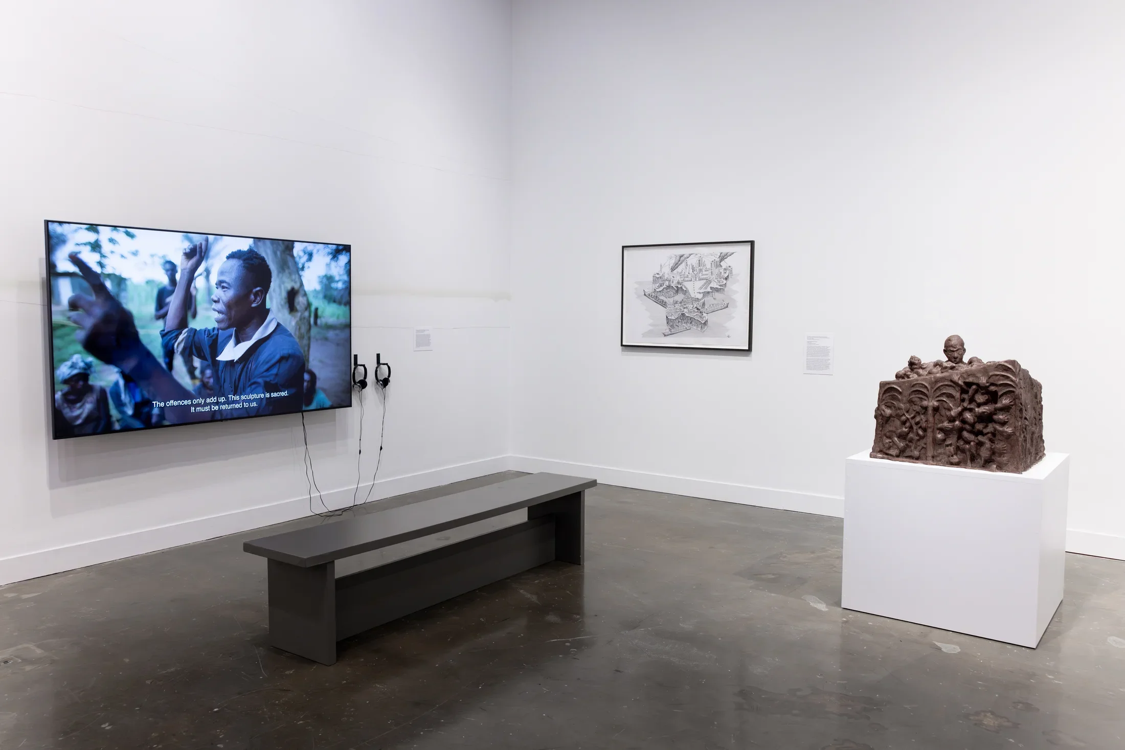 A group of artworks—including a flatscreen monitor, a wooden sculpture on a white pedestal, and grayscale drawing—occupy a corner of a gallery. The screen displays a video of a person speaking, with subtitles reading 'The offences only add up. The sculpture is sacred. It must be returned to us.' 