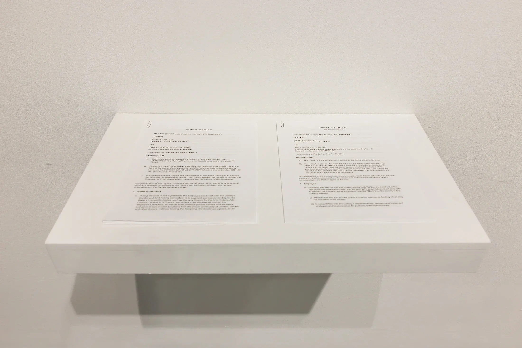 Two printed documents, appearing to be contracts, are displayed side-by-side on a white shelf against a white wall. The left is labeled “Contract for Services” and the right “FOREST CITY GALLERY Exhibition Contract.” The visible text includes dates, the names of parties to the contracts, and other legal text.