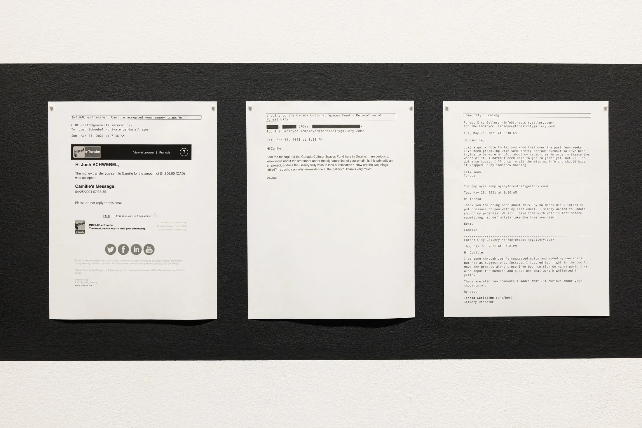 Three printed emails are displayed side-by-side on a white wall, showing correspondence related to a money transfer to someone named Camille; a confused inquiry from a granting agency about an employee’s email signature; and the gallery director’s burnout. The emails are dated April and May 2021.