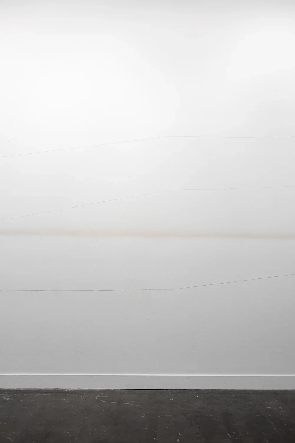 A white wall features three faint, thin horizontal lines that gradually crest upwards and downwards, as well as a more diffuse light brown or gray horizontal band across the middle. The floor in the foreground is dark gray.