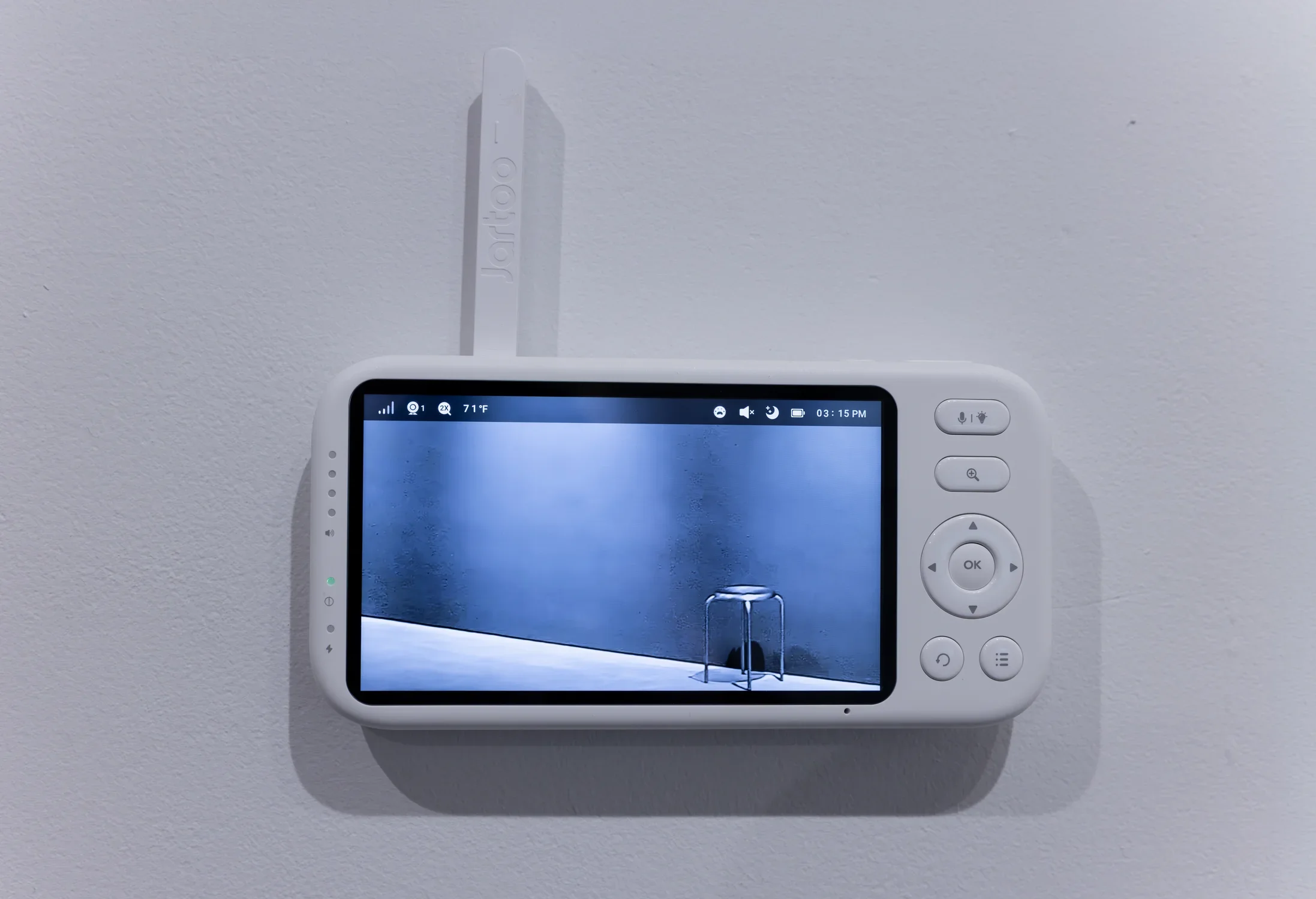 The viewing screen of a baby monitor—with navigation and menu buttons and a white antenna—is attached to a white wall. The feed on the screen shows a single metal stool against the background of dark gray walls and a sliver of concrete floor.
