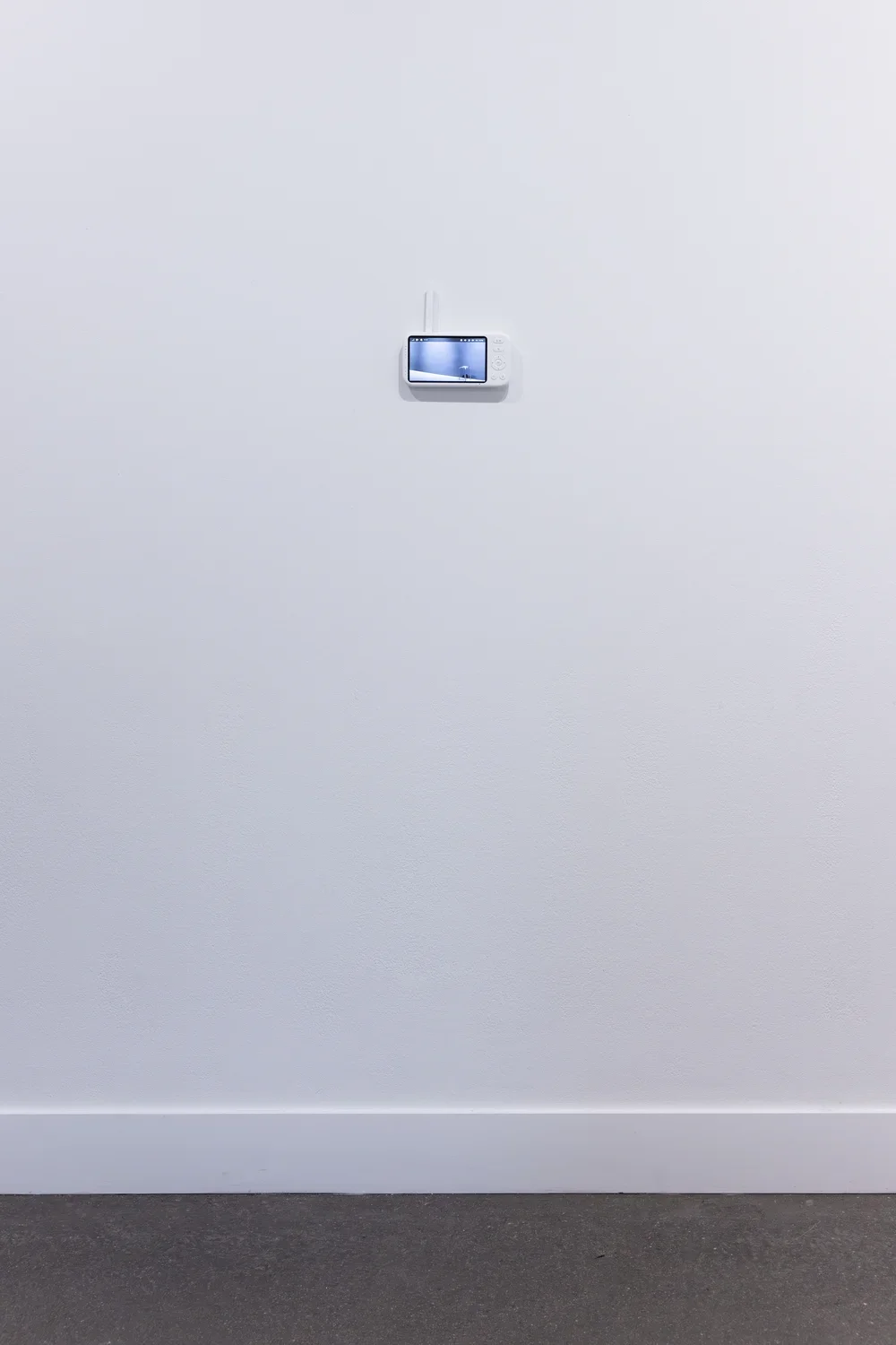The viewing screen of a baby monitor—with navigation and menu buttons and a white antenna—is attached to a white wall. It is dwarfed by an expanse of white wall and a sliver of gray floor. The feed on the screen shows a single metal stool.